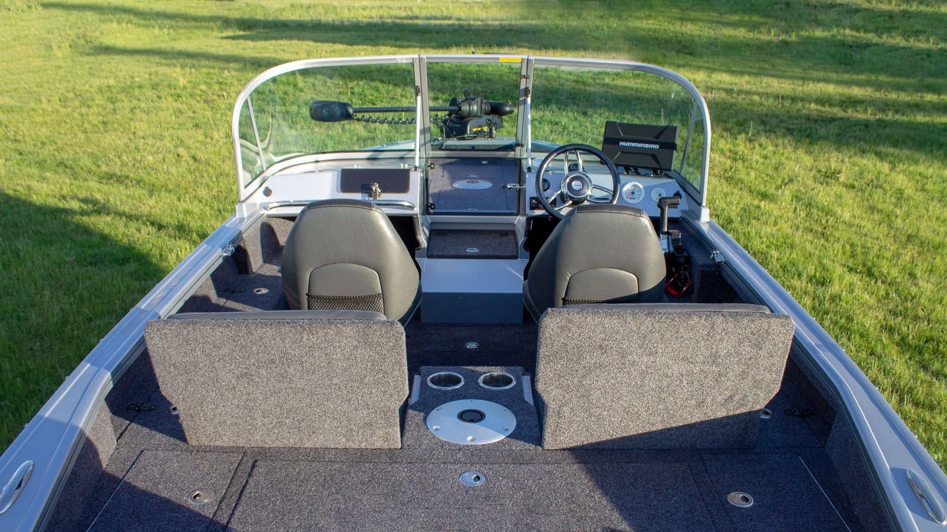 Inside view of a boat on green grass, showing two front seats, a steering wheel, a GPS display, and fishing accessories.