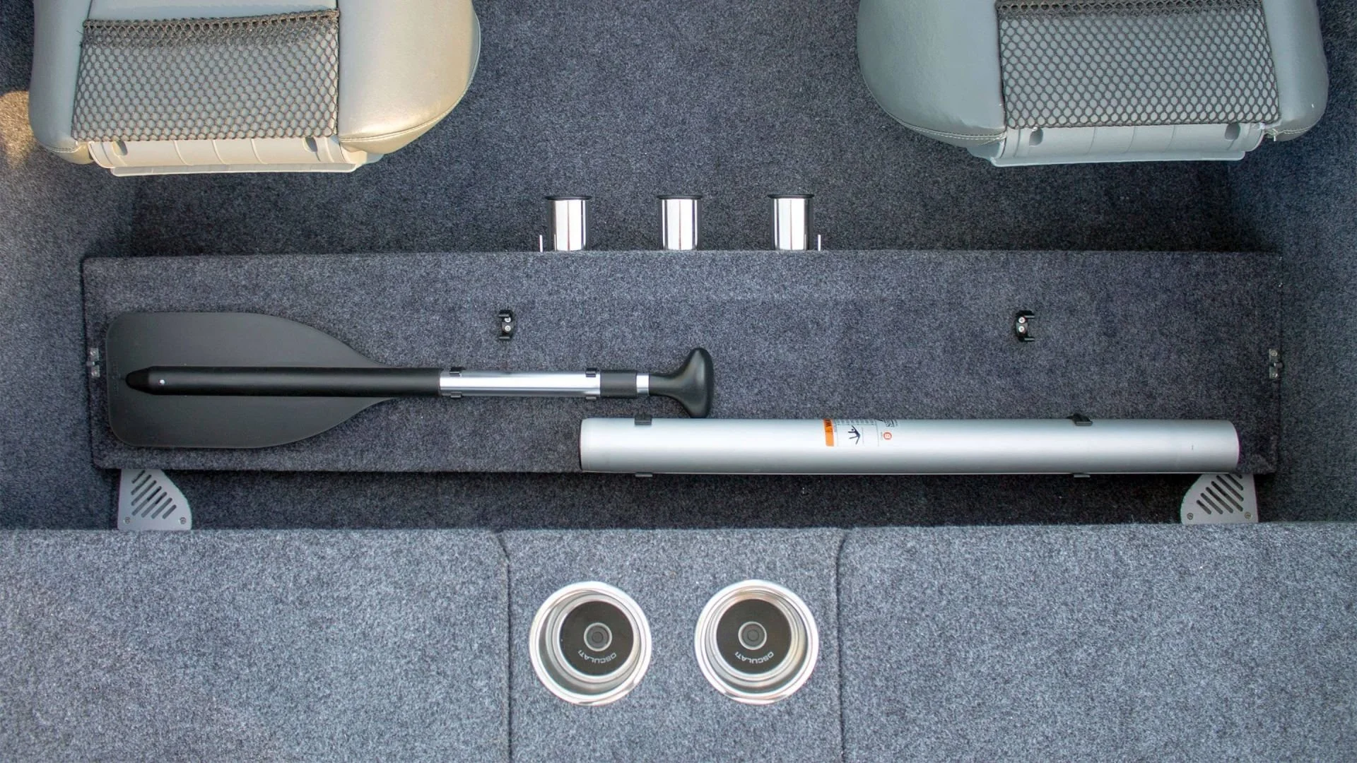 A boat's carpeted storage compartment containing a collapsible shovel, a silver cylindrical object, and four small cans or containers, with two beige seats in the background.