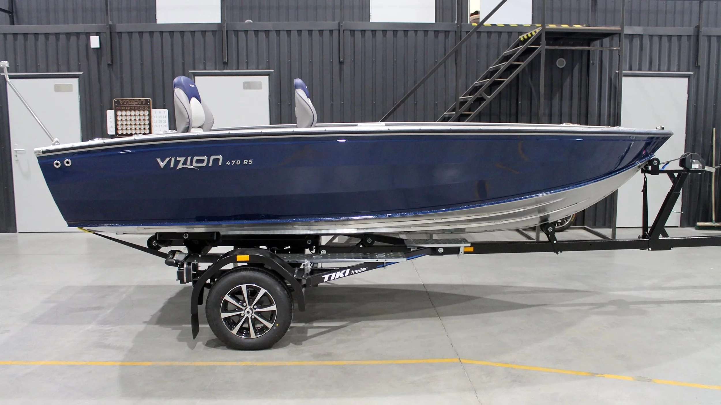 A blue summer boat labeled 'VIZION 470 RS' on a black trailer inside a warehouse with black walls and gray doors.