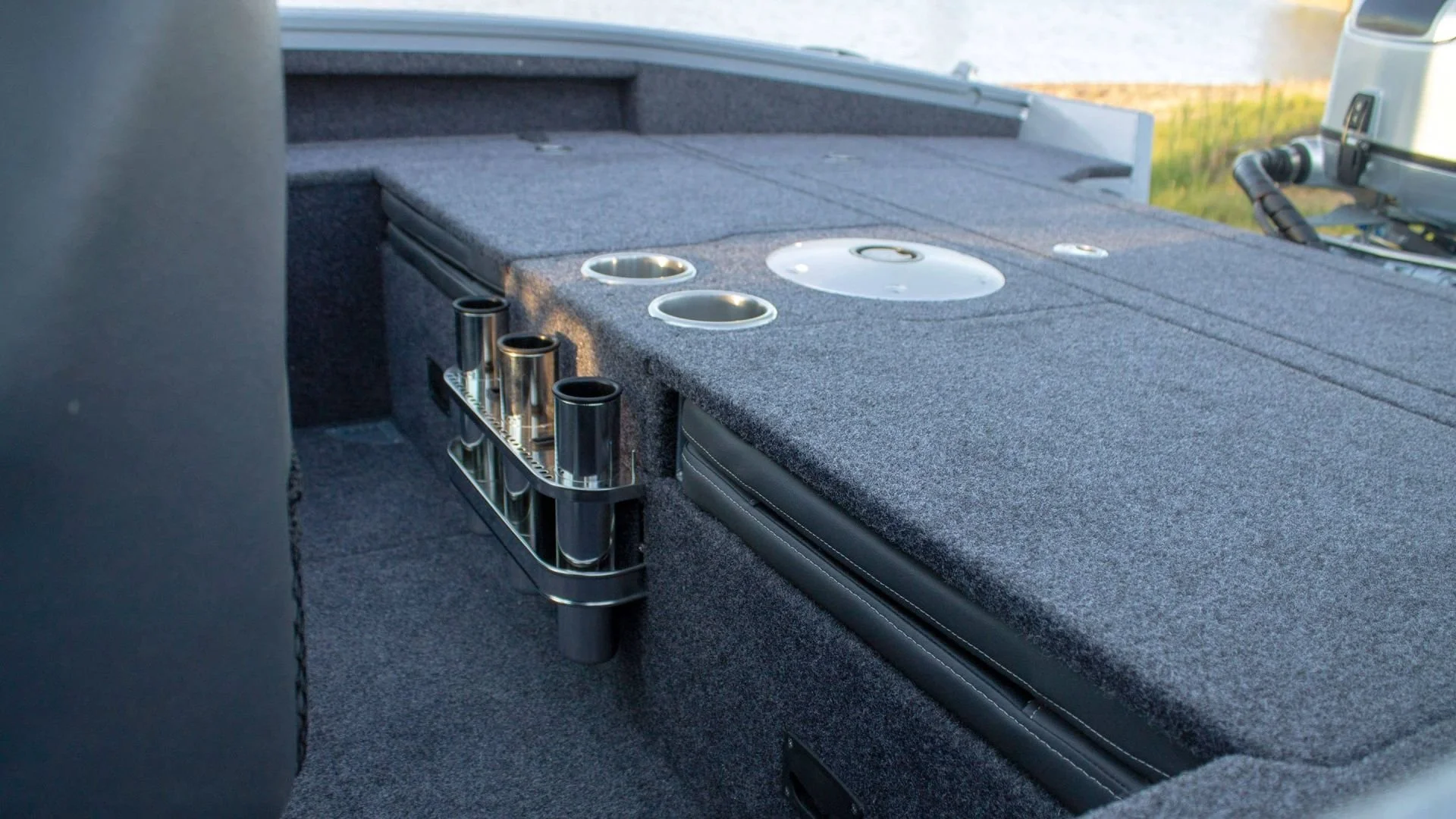 The image shows the interior of a boat with a gray carpeted surface, cup holders, and a metal rack holding black tubes or rods.