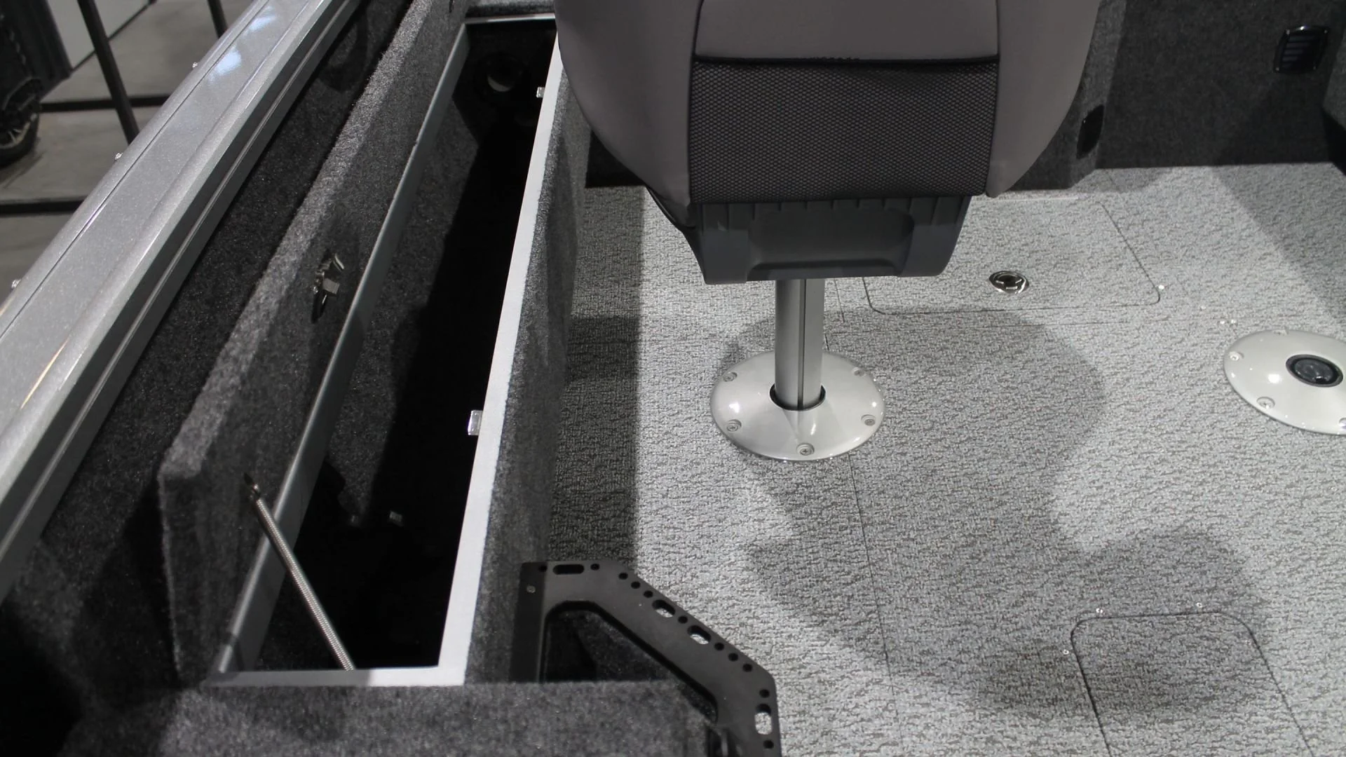Interior of a boat with a gray carpeted floor, a gray swivel chair, and an open storage compartment with foam padding and a metal latch.