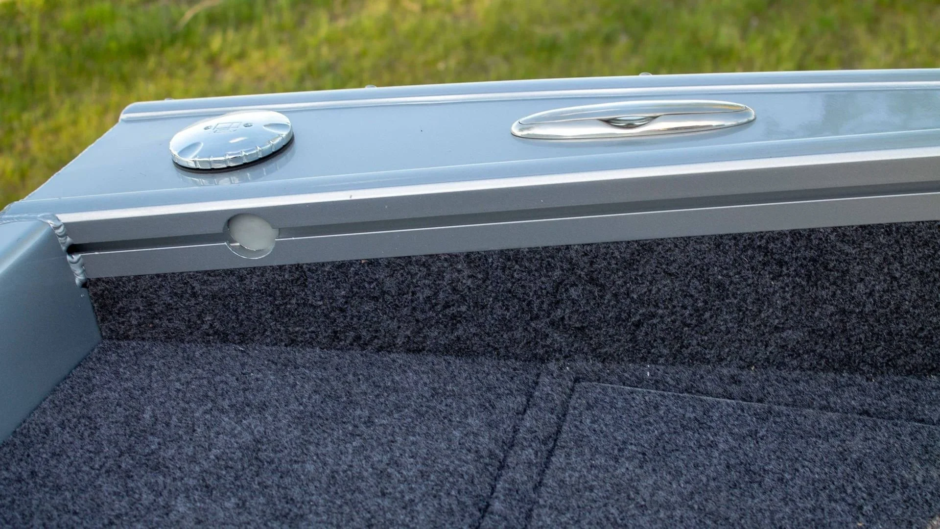 Close-up of the edge of a boat with a silver fuel cap, a silver handle, and a foam gasket, set on a grassy area.