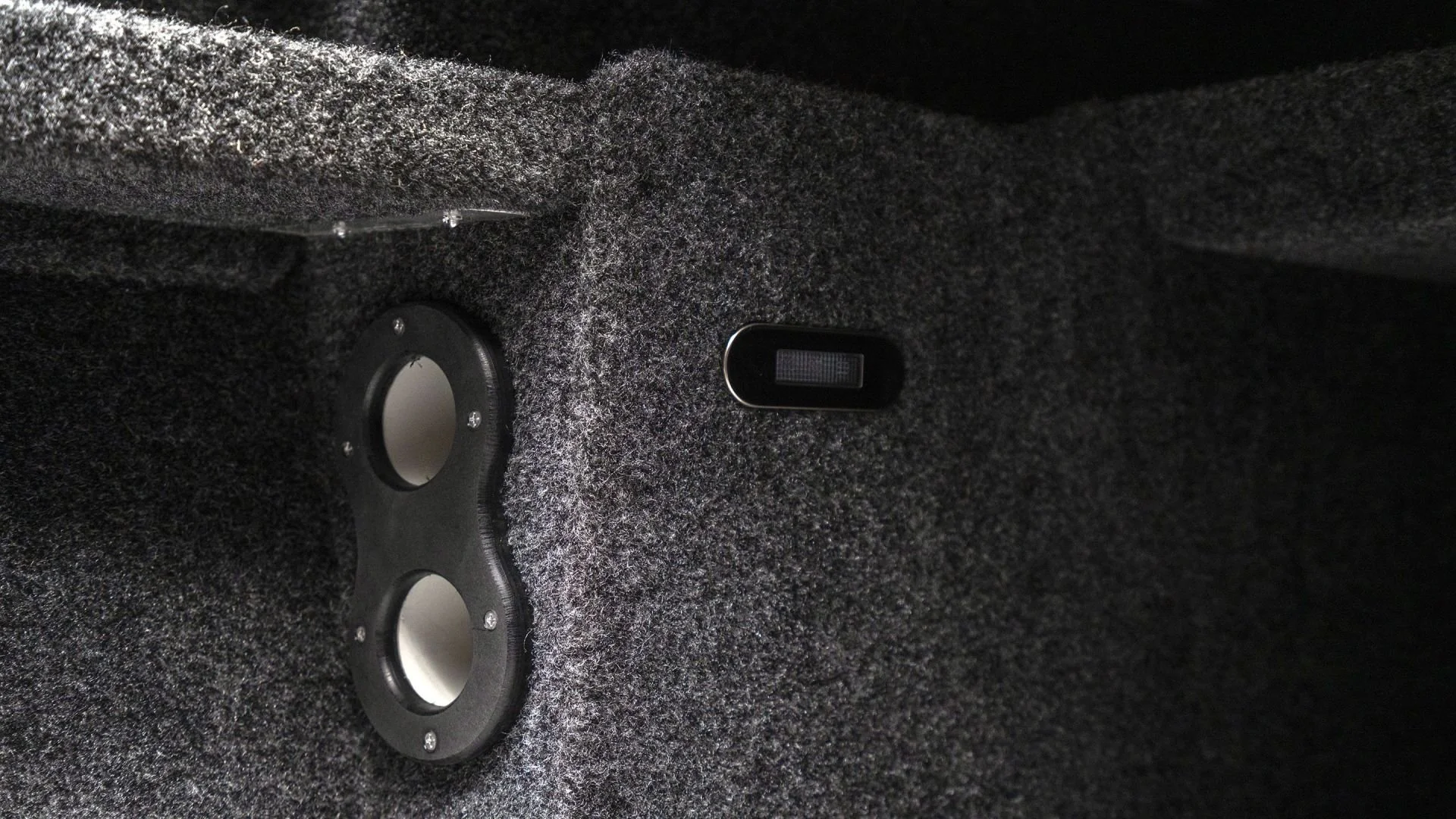 Close-up of a car's trunk interior with two circular subwoofers installed and a small black switch or control mounted on the side.