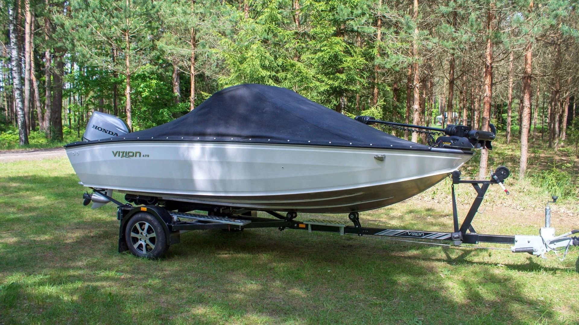 A boat on a trailer parked on grass near a forested area with tall trees and green foliage.