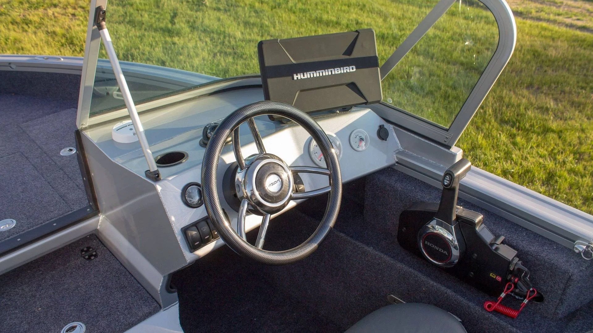 Interior of a small boat with a steering wheel, electronic equipment, and a mounted GPS device, with a grassy field visible through the windshield.