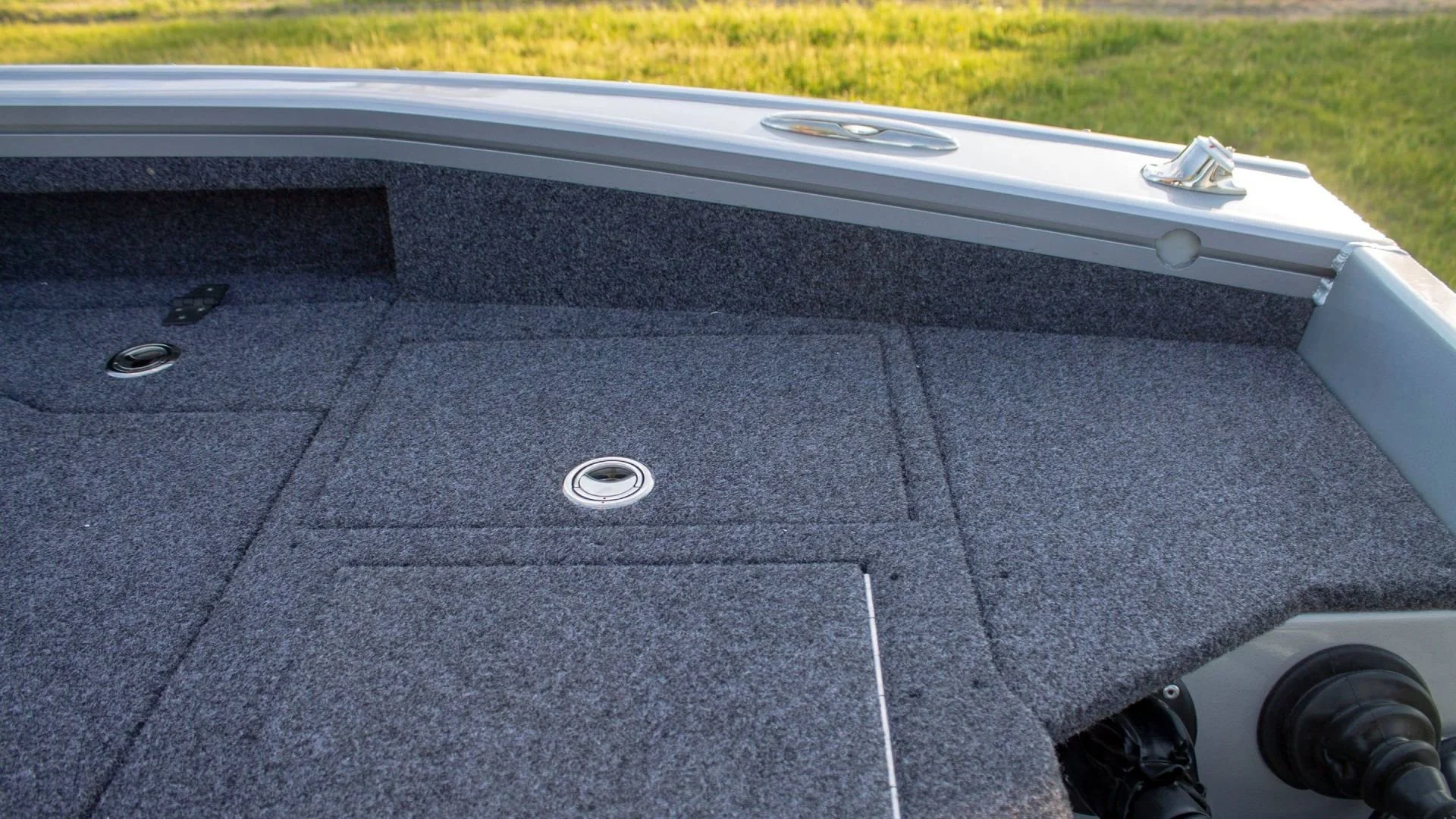 Empty fishing boat deck with carpeted surface and a few small compartments, surrounded by green grass.