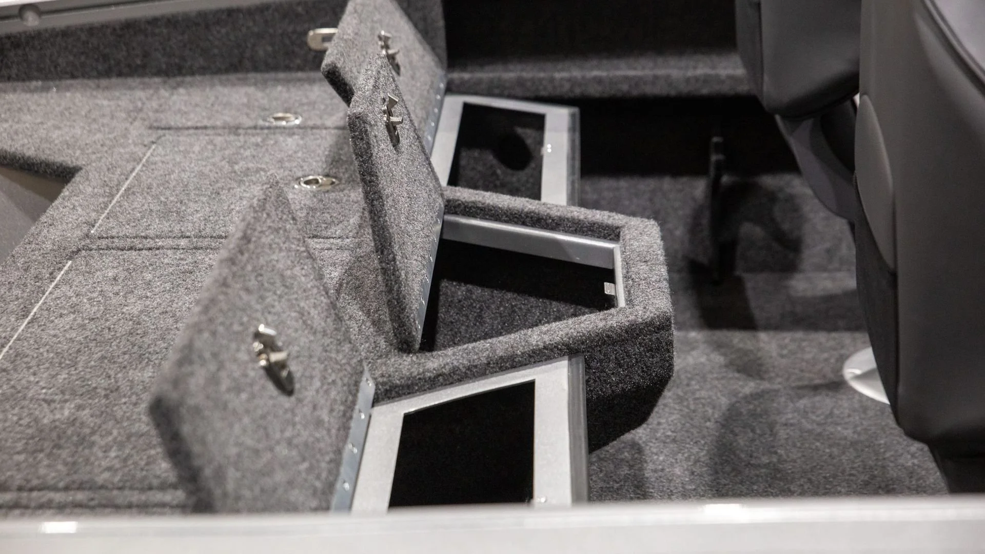 The interior of a vehicle showing a section of gray carpeted storage compartments and frames for mounting electronic devices or screens.