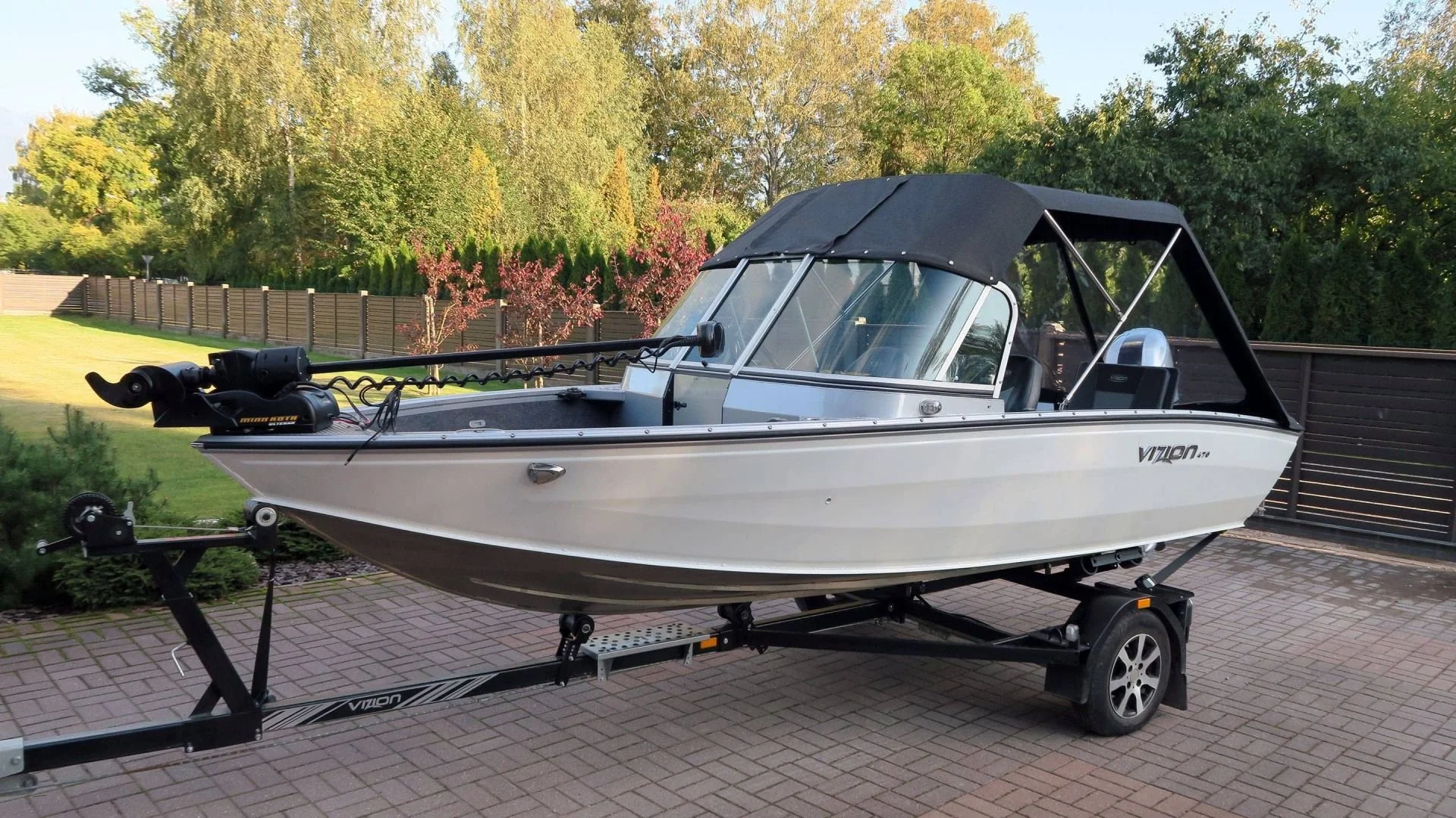 A small white motorboat on a black trailer parked on a brick driveway, with a black canopy, windshield, and a trolling motor at the bow in a yard with trees and a fence in the background.