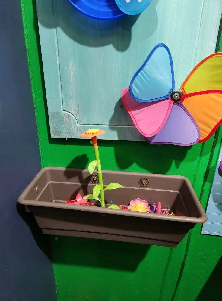 A brown plastic flower box sticking out from a wall with plastic flowers growing from it. On the wall are colorful flower decorations.