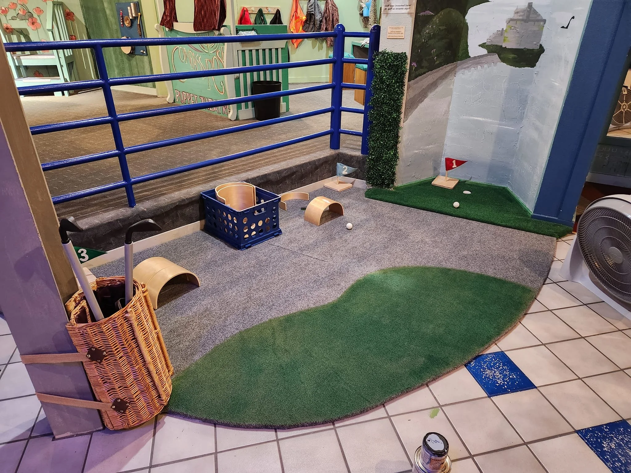 Indoor miniature putting green with a painting of the Loch Ness on the far wall. The putting green backs up against a railing.