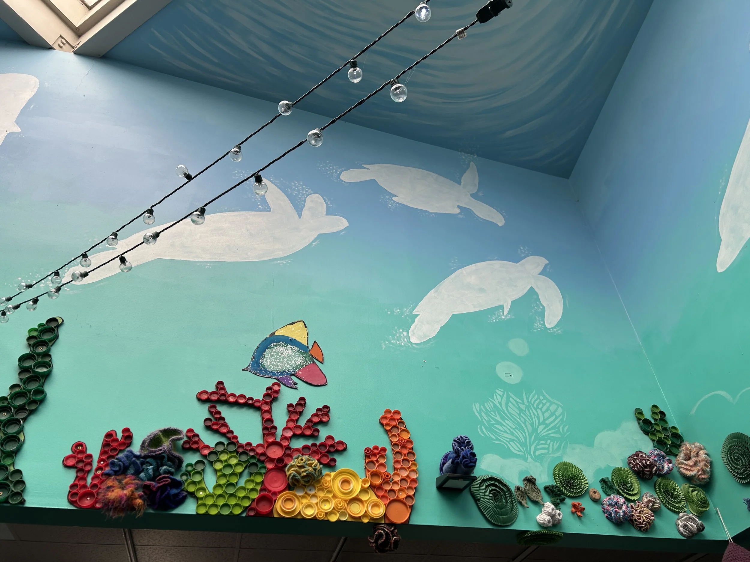 Ocean-themed wall art, including painted sea turtles and colorful coral made from recycled bottle caps. A skylight can be seen in the upper corner, and string lights hang from somewhere offscreen.