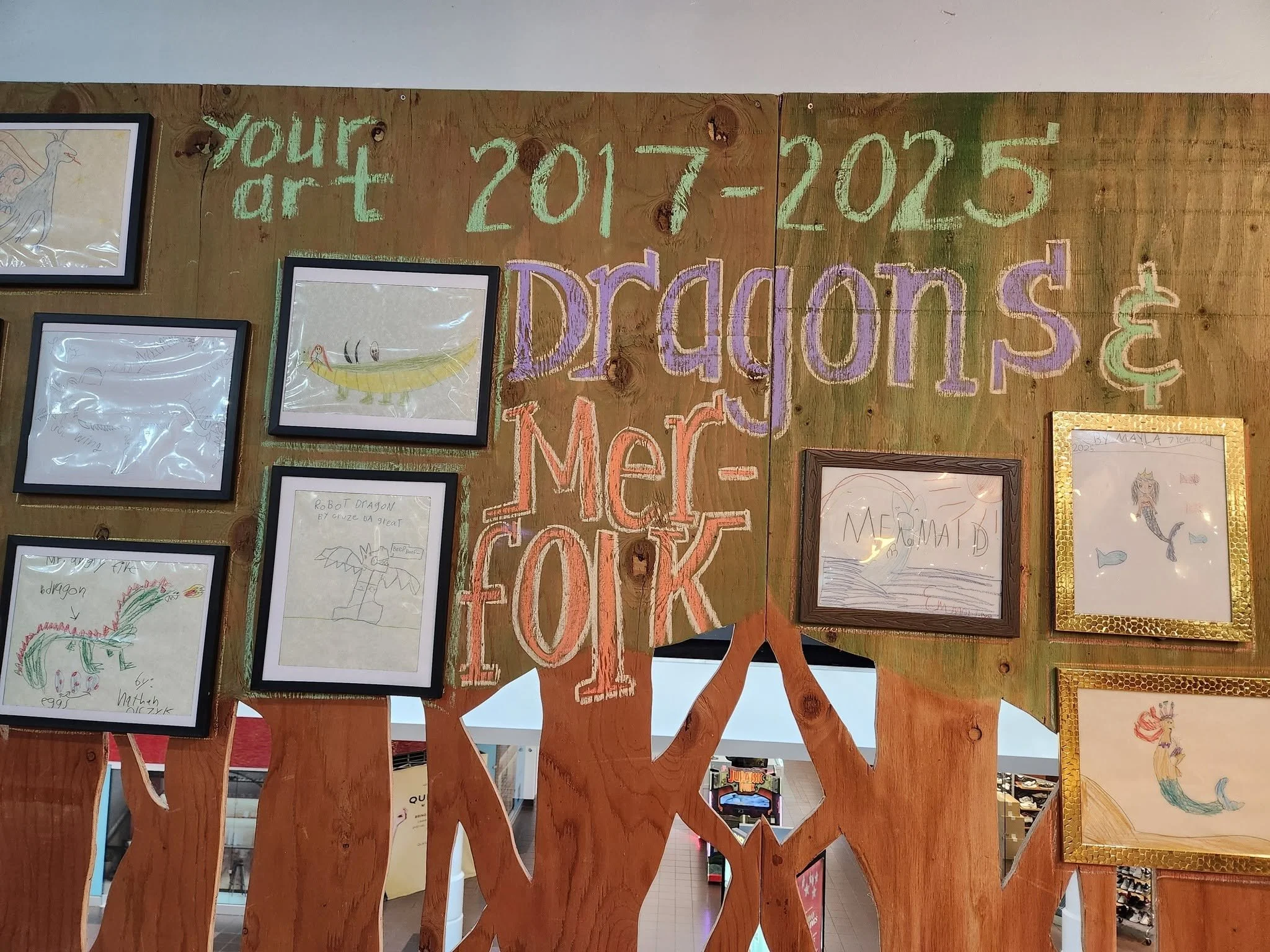 A tree line made out of wood lines a wall. On the wall, chalk writing describes dragon and merfolk artwork from 2017-2025. Next to the writing are frames holding pictures of the dragon and merfolk artwork.
