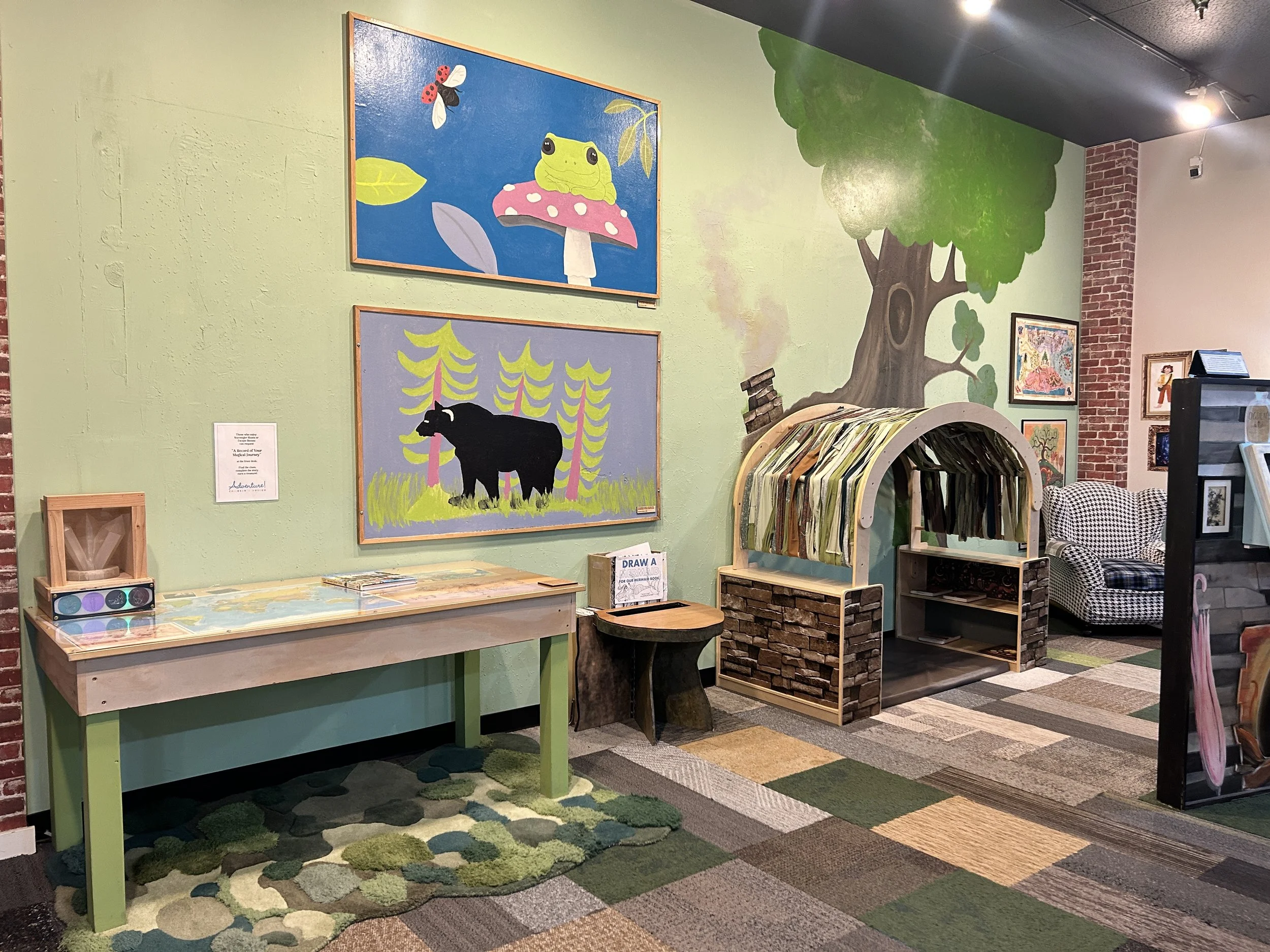 The entrance to the Magic and Mystery Exhibit, brightly lit and colorful. On one wall, two paintings of forest creatures are hung. There's a hobbit-like hut structure for children to climb into against one wall, with a painted tree coming out of it.