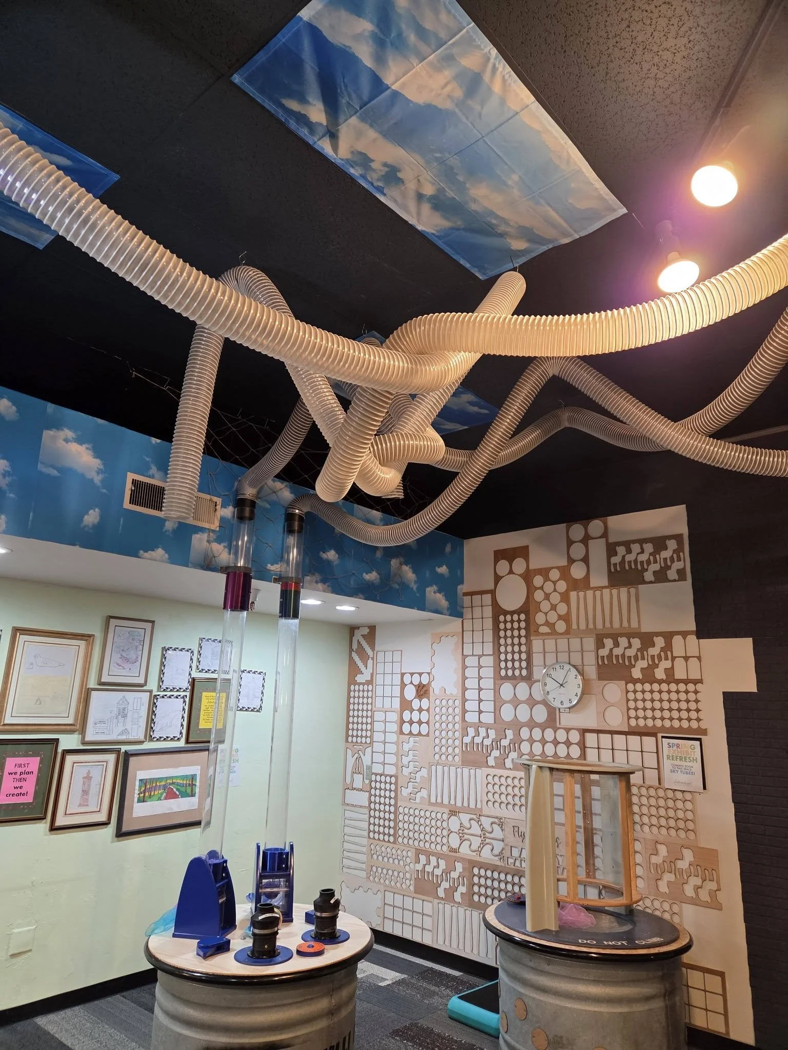 A room with a dark gray ceiling and PVC tubes hanging from the ceiling, using airflow to shoot scarves through the tubes. On the far wall, framed photos of blueprints hang.