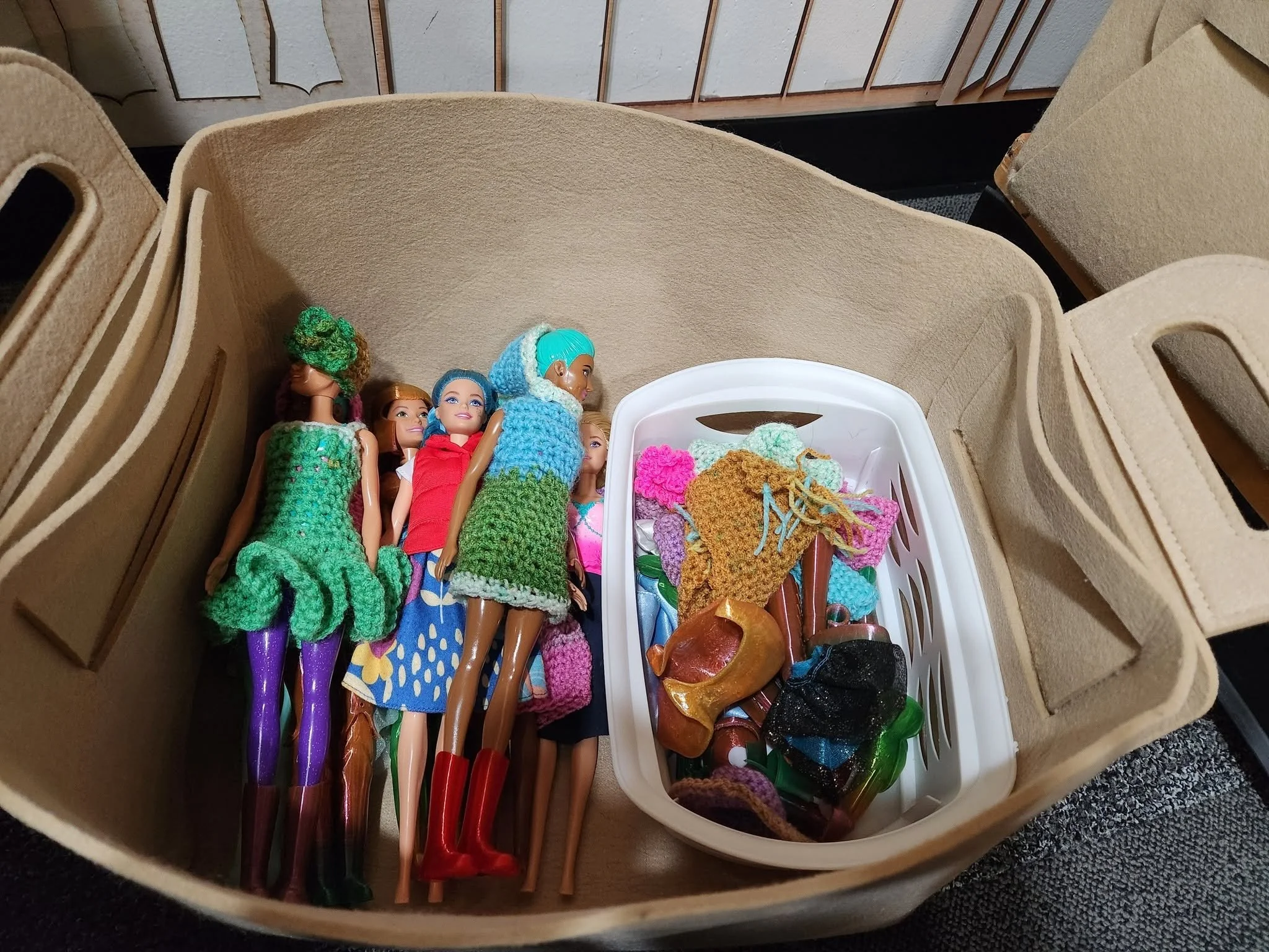 A soft tan-colored bin holding a small white plastic bin full of Barbie clothes inside, as well as four or five Barbies dressed in various clothing.