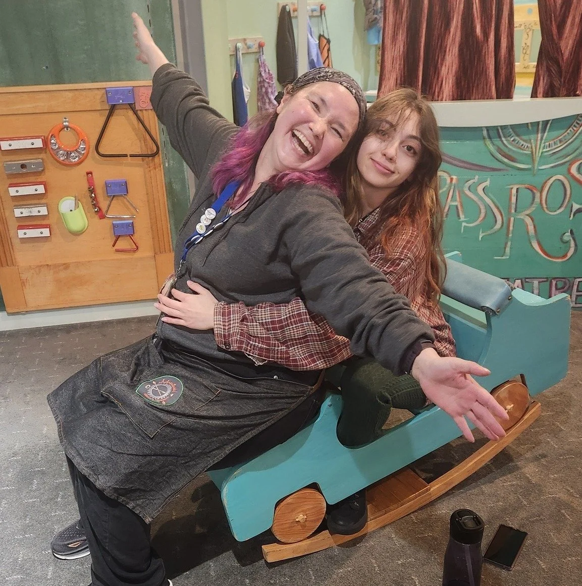 Two staff members sitting on a wooden toy car painted blue. One staff member has her arms wrapped around the other, while the other holds her arms out in an airplane-like pose. They both smile to the camera.