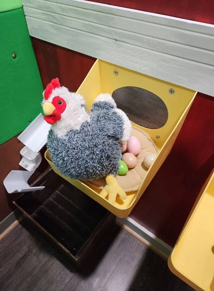 A plush rooster sitting on a plastic yellow chicken coop. Peeking out from underneath the rooster are plastic eggs.