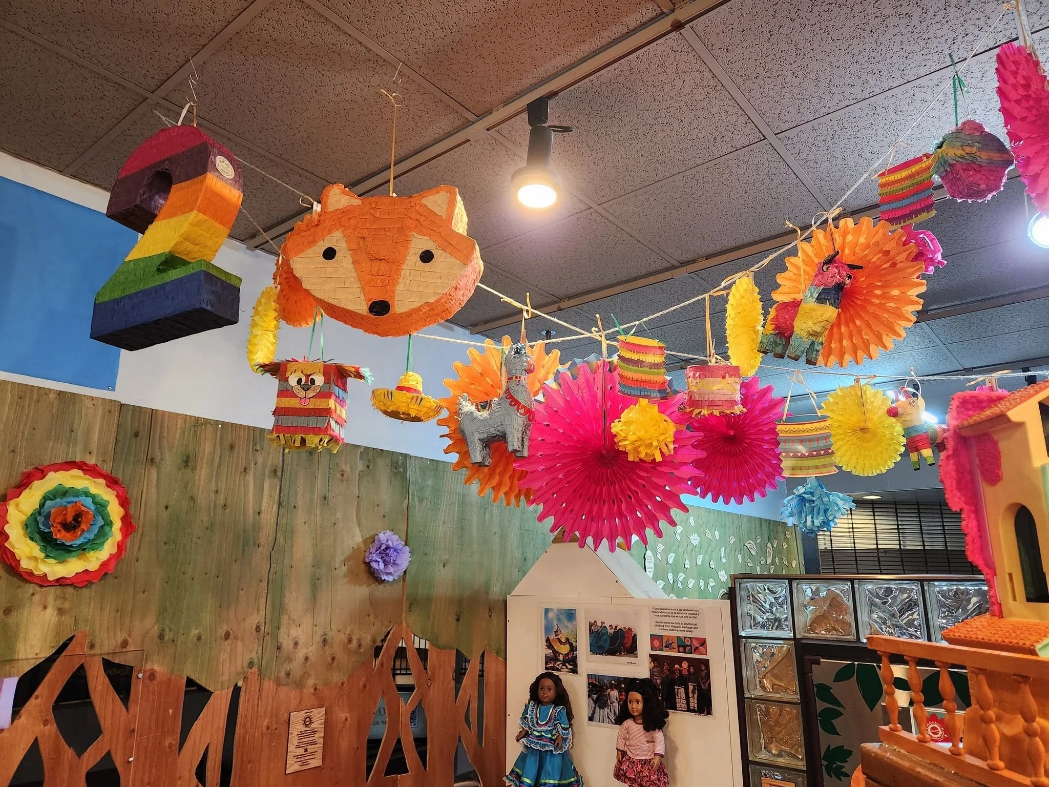 Colorful pinatas hang from the ceiling of the Hispana Herencia exhibit. On the far wall is a tree line made out of wood.