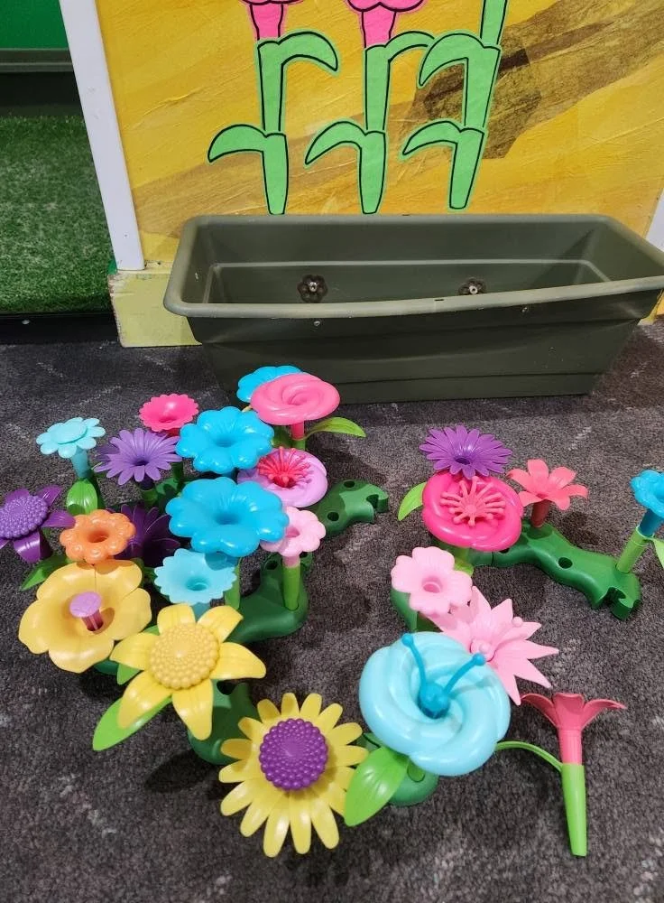 Plastic flowers lined up on the floor in front of the Flower Garden exhibit. In the background, a plastic flower bed is attached to a wall with paintings of flowers on it.