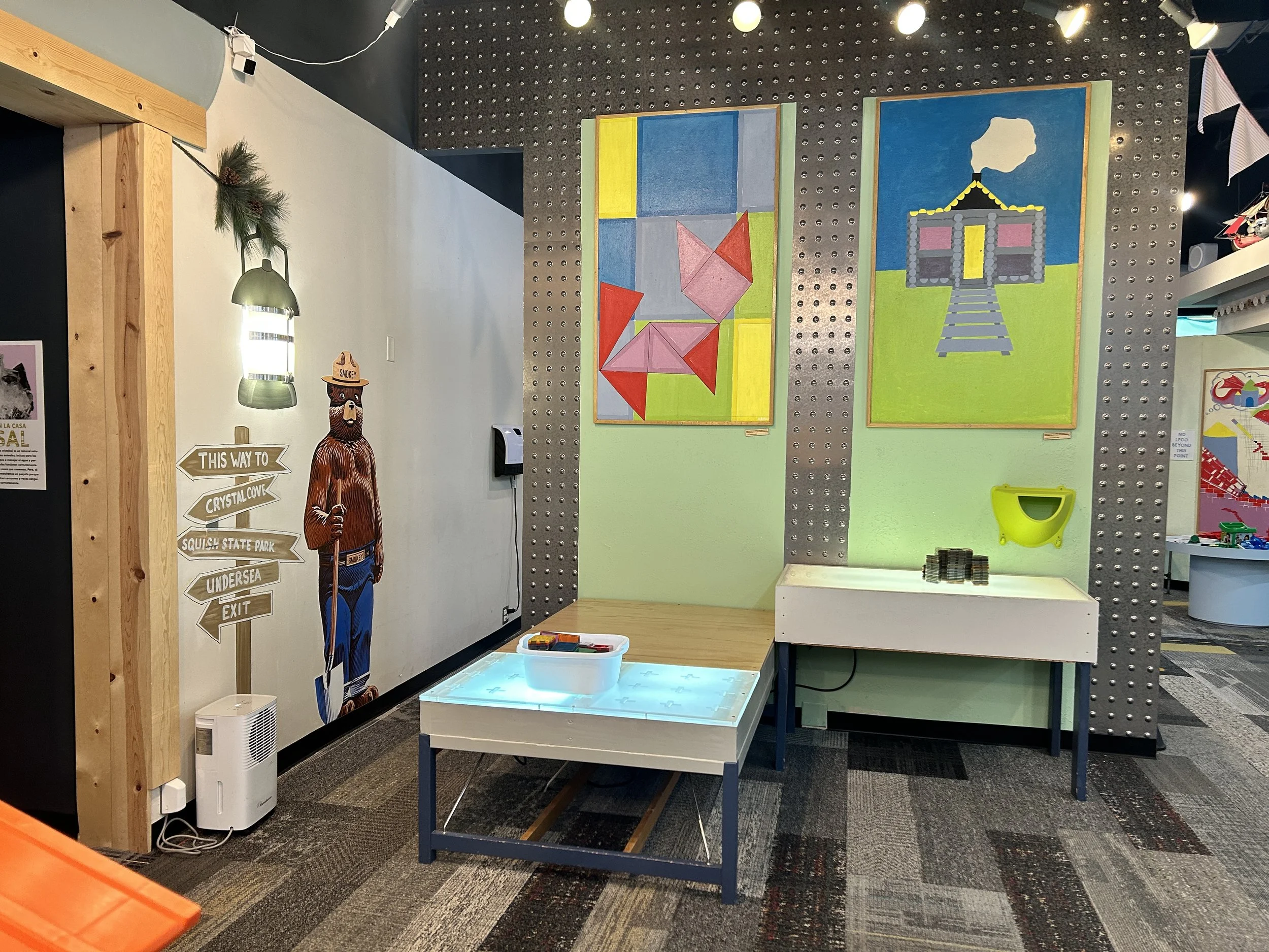 Two light tables turned on to illuminate Magnetic Tiles. On the wall above, painted abstract art is hung. On the wall to the left, there's a poster of Smokey the Bear, a lamp, and signs pointing to different directions.