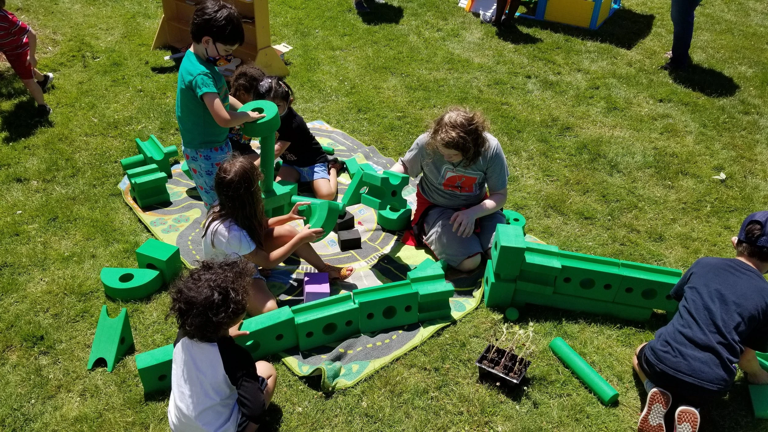 Children playing outside in the grass with green foam blocks of various sizes.