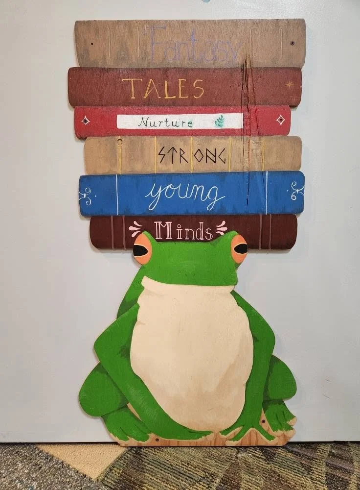 A painted mural of a green frog with books on its head. The books have their spines facing outward, with "Fantasy books nurture strong young minds" written one word per book.