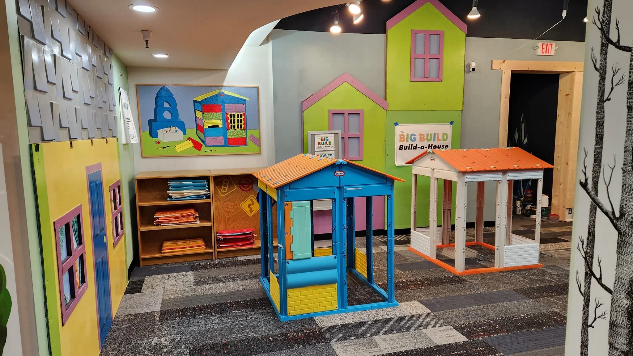 Colorful and bright play area with two house structures about 4 feet in height. Panels for the houses sit on a nearby shelf, and the wall is decorated with colorfully painted houses out of cardboard.