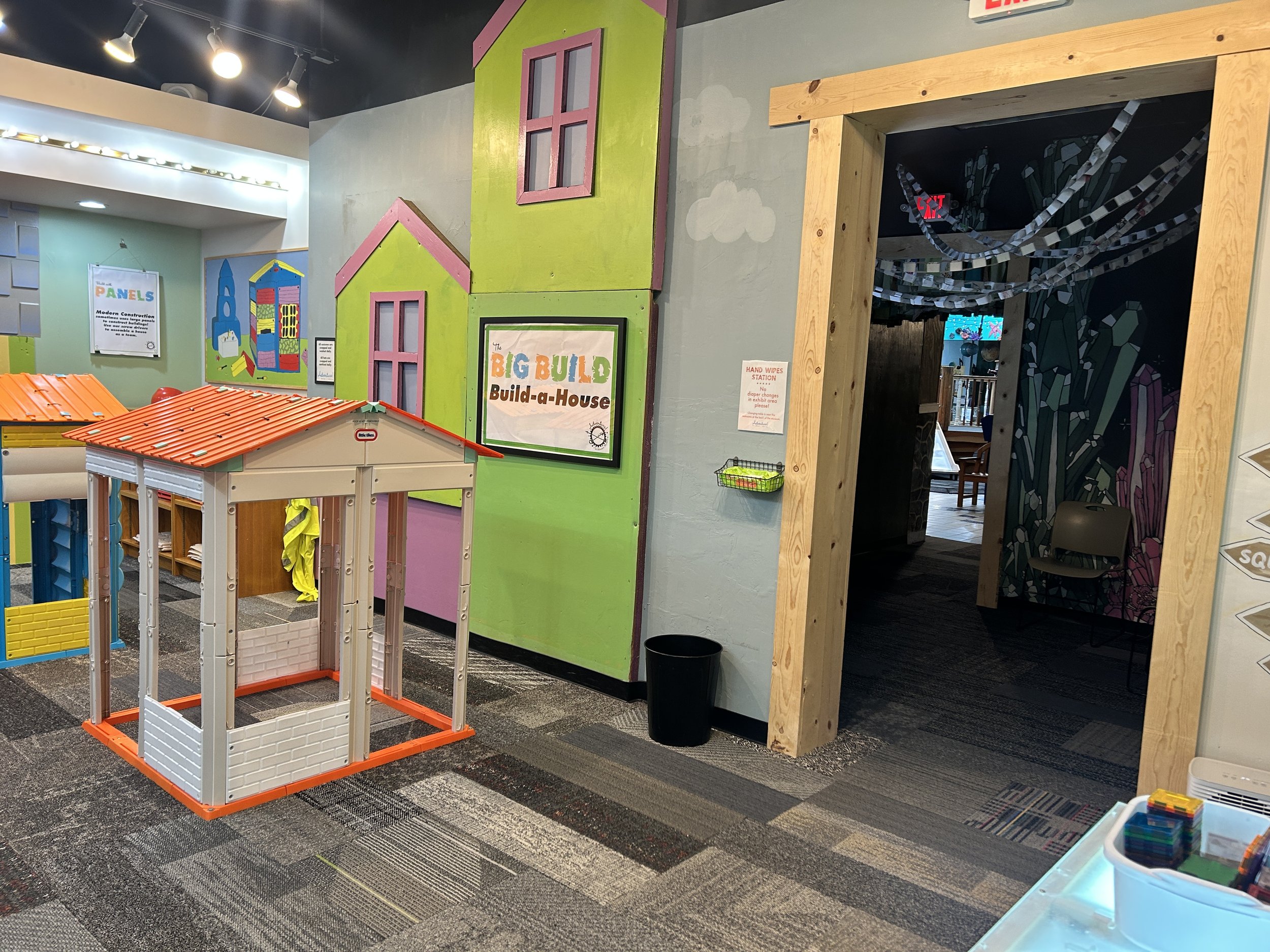 An entryway to a room with the Build-a-House structures and build tables. The wall to the left of the entryway is covered in cardboard cut out and decorated to look like houses.