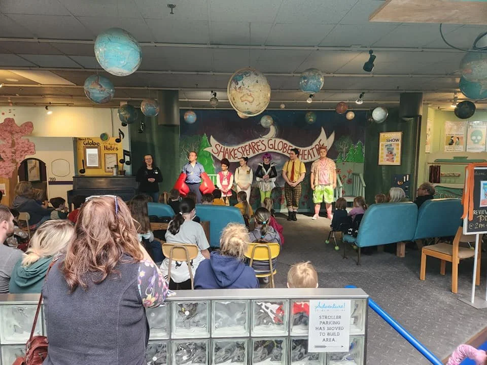Families watching a performance at the Shakespeare's Globe Theatre, an open space with globes hanging from the ceiling.