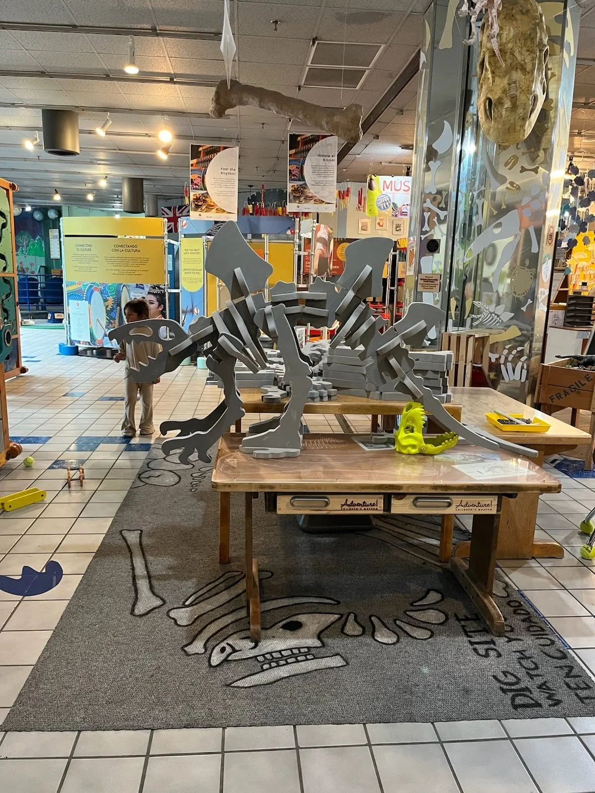 A view of the Dino Dig Site exhibit, featuring three tables side by side. One of the tables, the longest, has giant gray foam blocks on it for building a dinosaur skeleton. On one of the other tables sits a dinosaur made from these foam blocks.