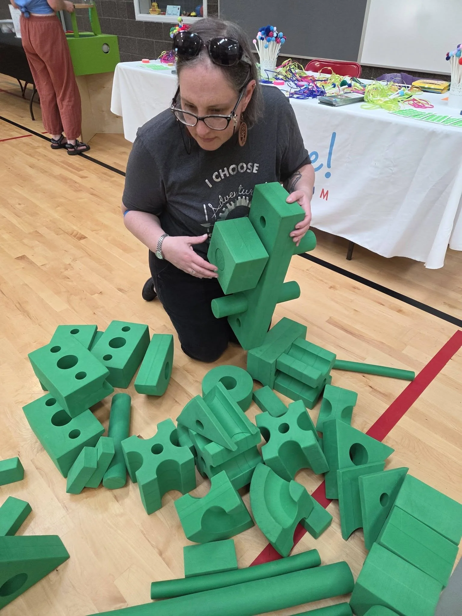A woman kneeling on a gym floor playing with green foam blocks. The green foam blocks are different shapes and sizes.
