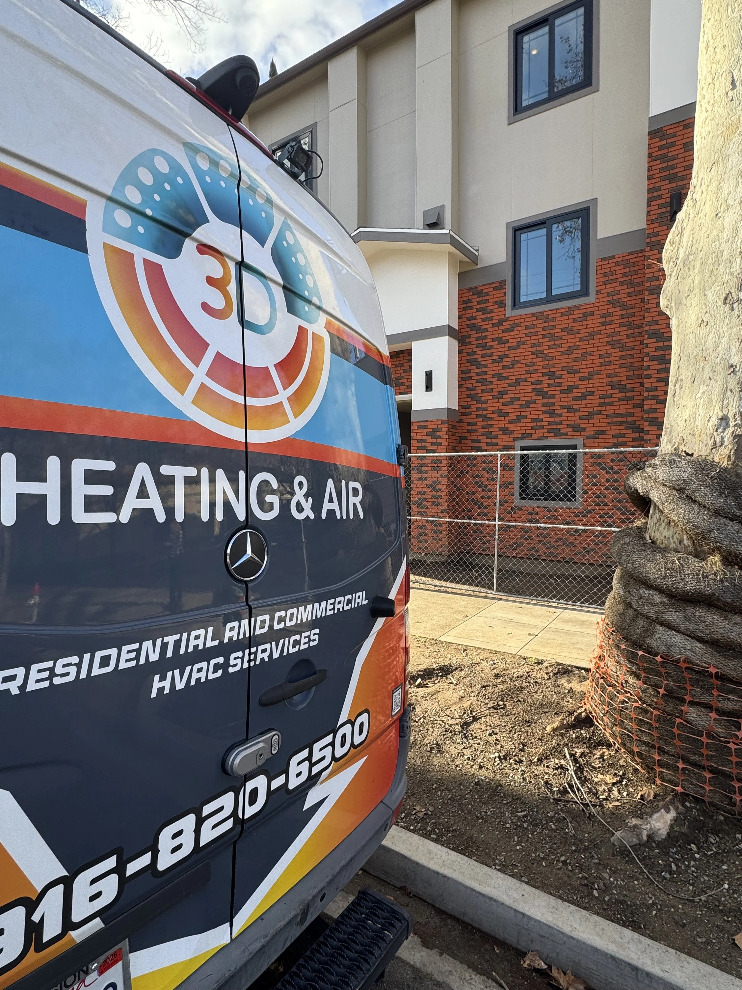 A service van for heating and air conditioning parked in front of a residential building with a tree and a construction material wrapped in orange mesh.