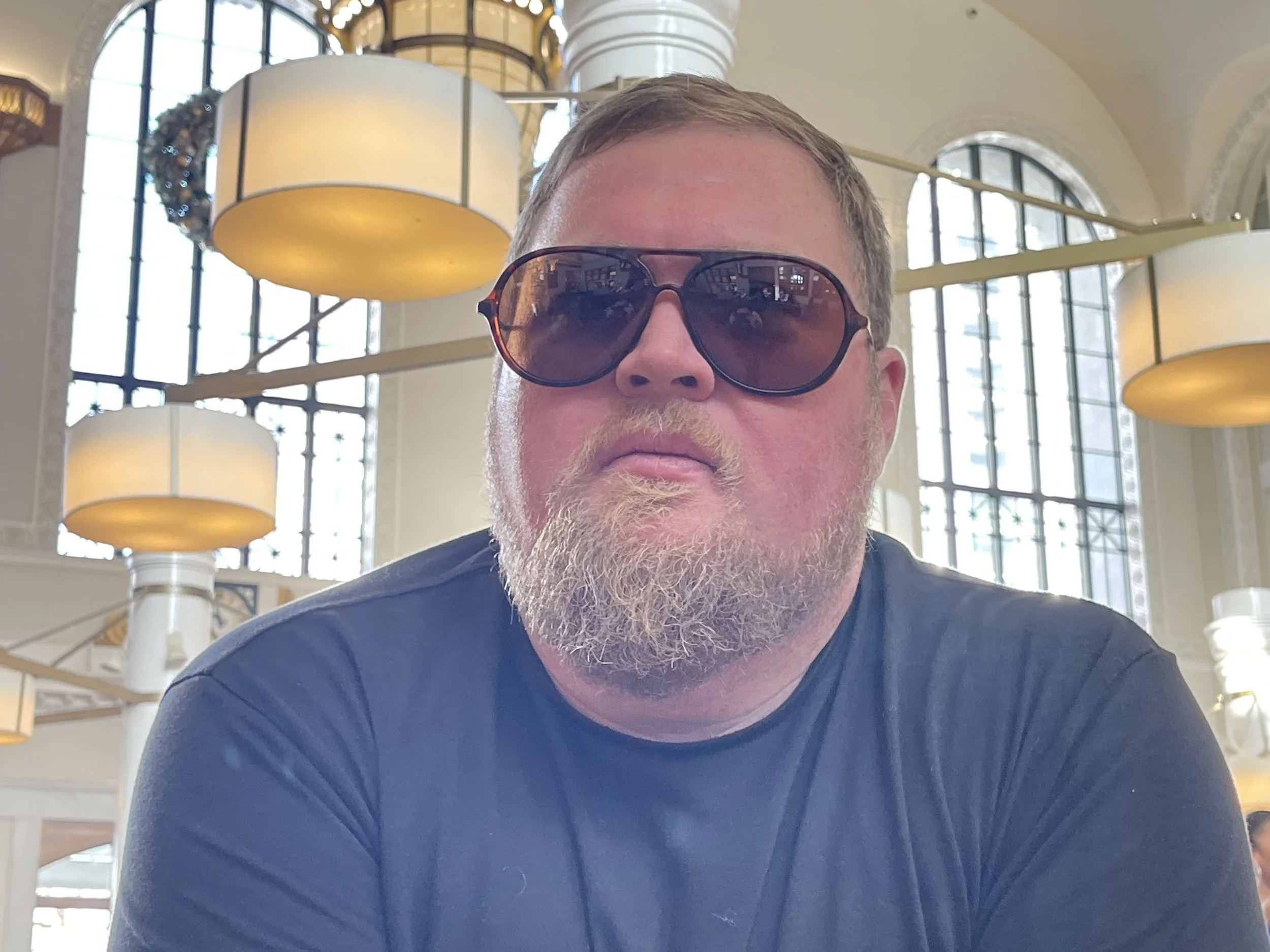 A bearded man wearing sunglasses and a black shirt sitting indoors with large windows and hanging lamps in the background.