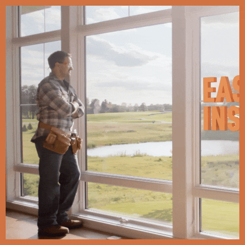 A man standing inside by large windows, looking out at a grassy landscape with a river and trees, with an orange sign that reads 'EAST INS' partially visible.