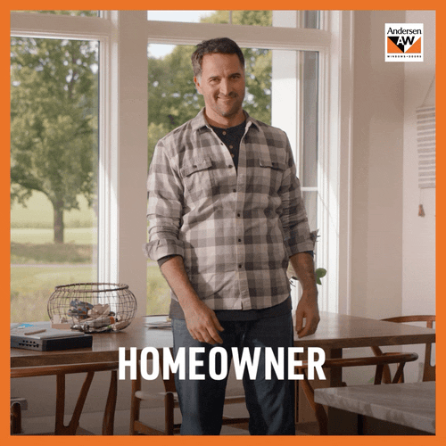 A man standing in a dining room with large windows, smiling, wearing a plaid shirt and jeans.