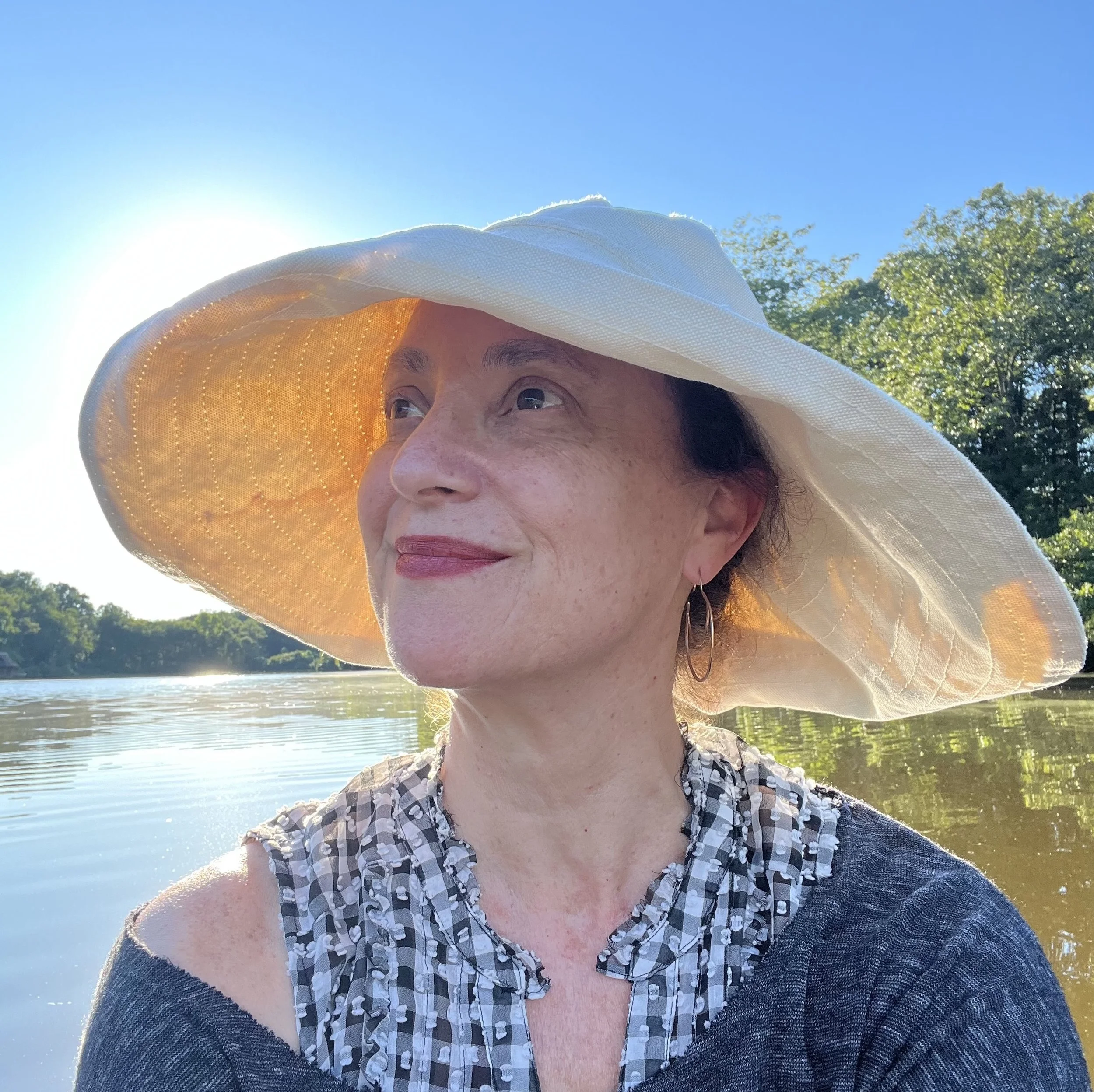 Victoria wearing a large white sun hat and a patterned top sitting by a river on a sunny day, with trees in the background.