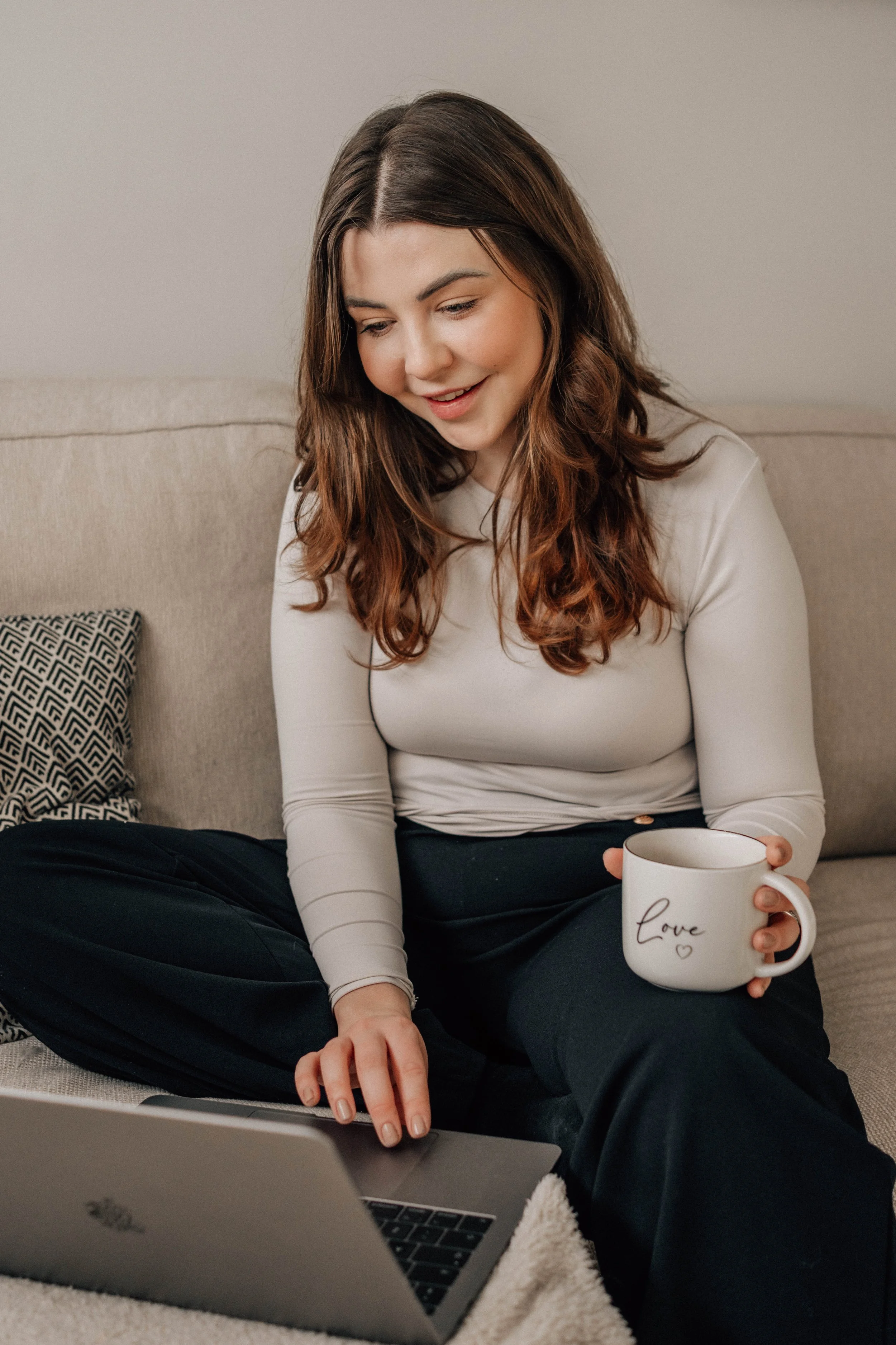 A woman with long brown hair, wearing a beige long-sleeve shirt and black pants, sitting on a beige couch with a patterned pillow, using a laptop. She is holding a white mug with the word 'Love' written on it.