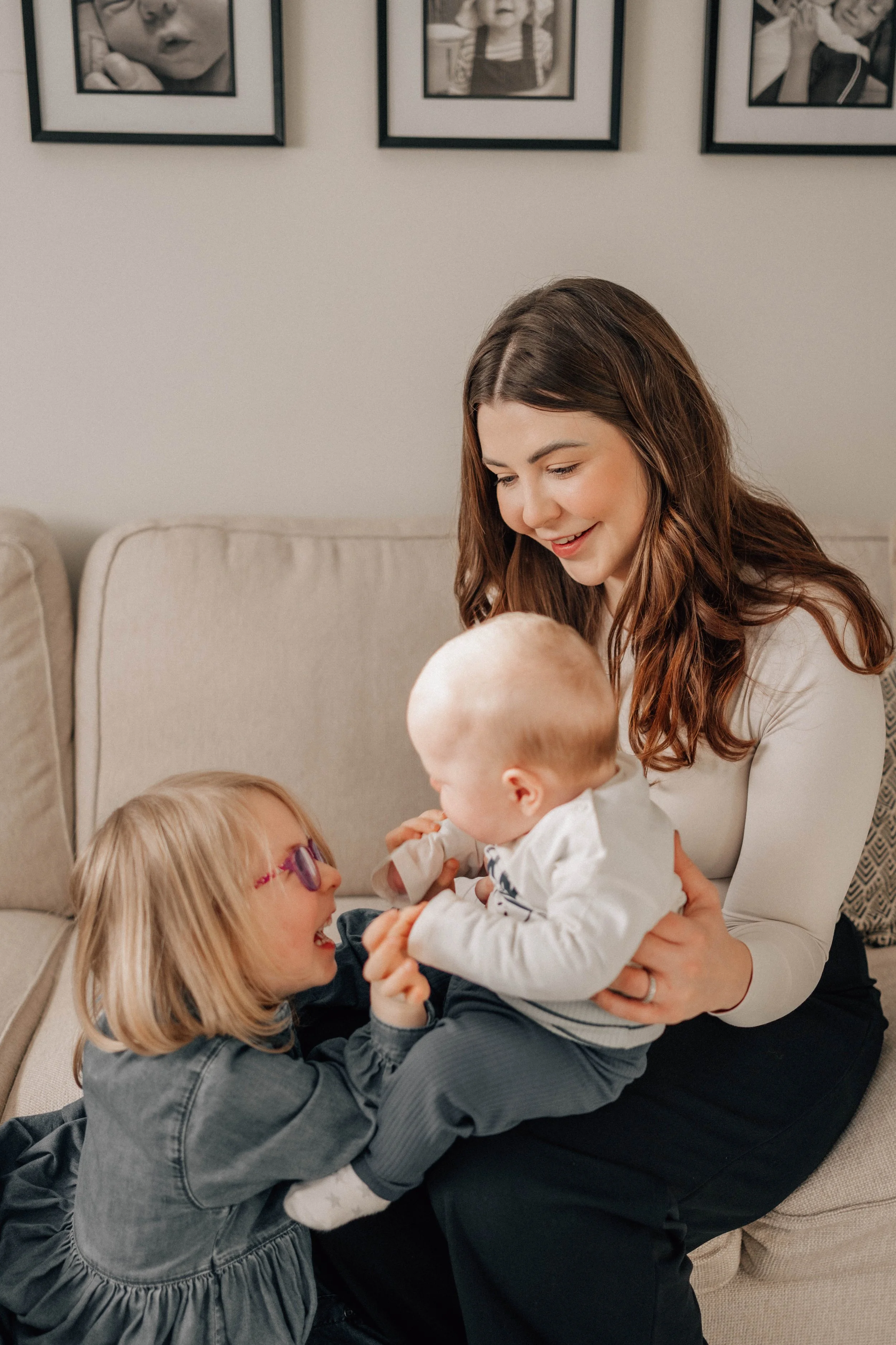 A woman sitting on a beige couch holding a baby, while a young girl in glasses and a dark jacket playfully pulls at the baby's hands, all smiling and enjoying a playful moment.