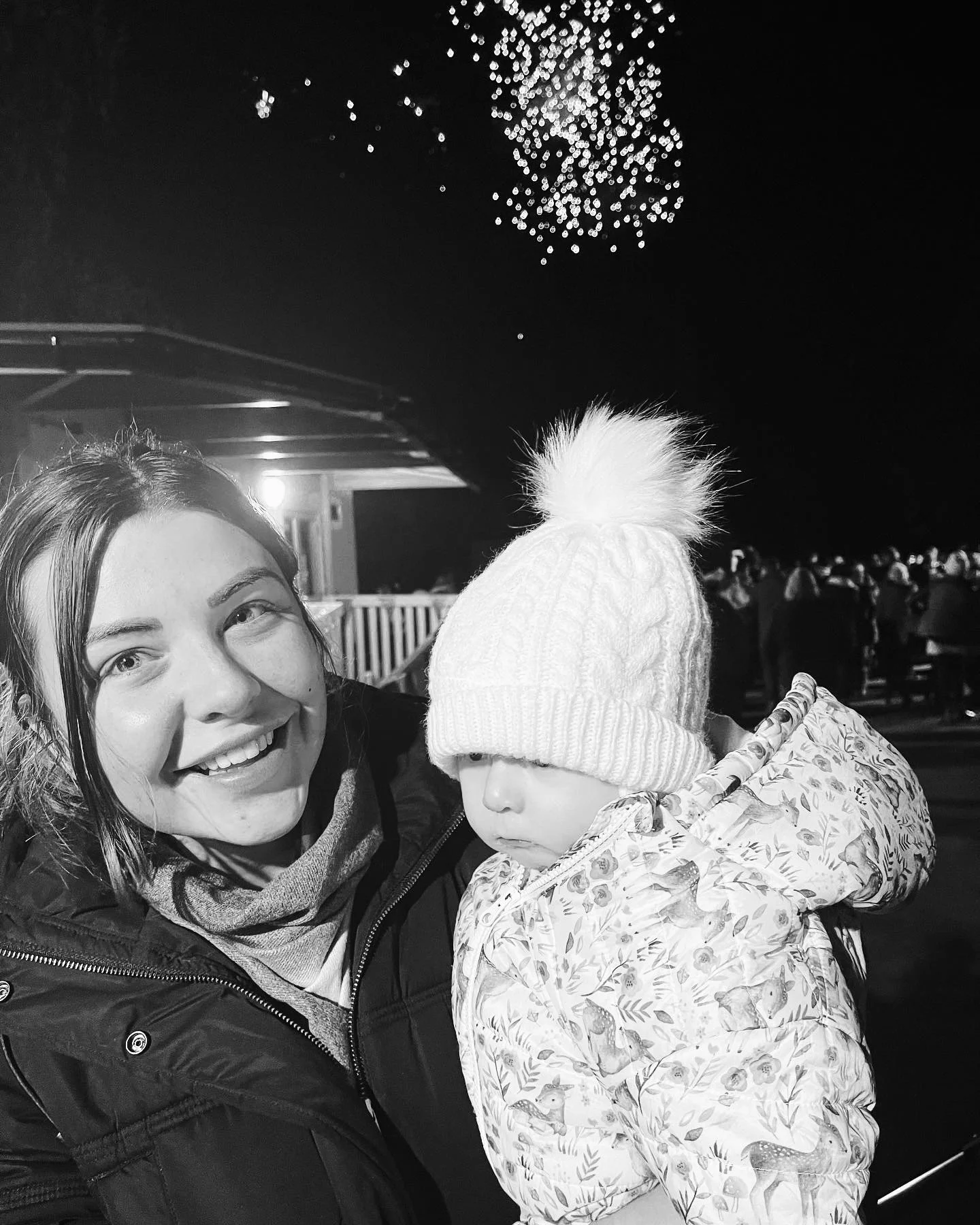 A woman smiling and taking a selfie with a young child wearing a winter hat at an outdoor event at night with fireworks in the sky.