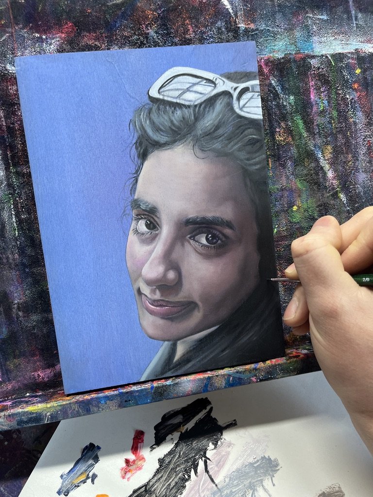 A person is painting a realistic portrait of a young woman on a blue canvas, with sunglasses resting on her head and a slight smile.