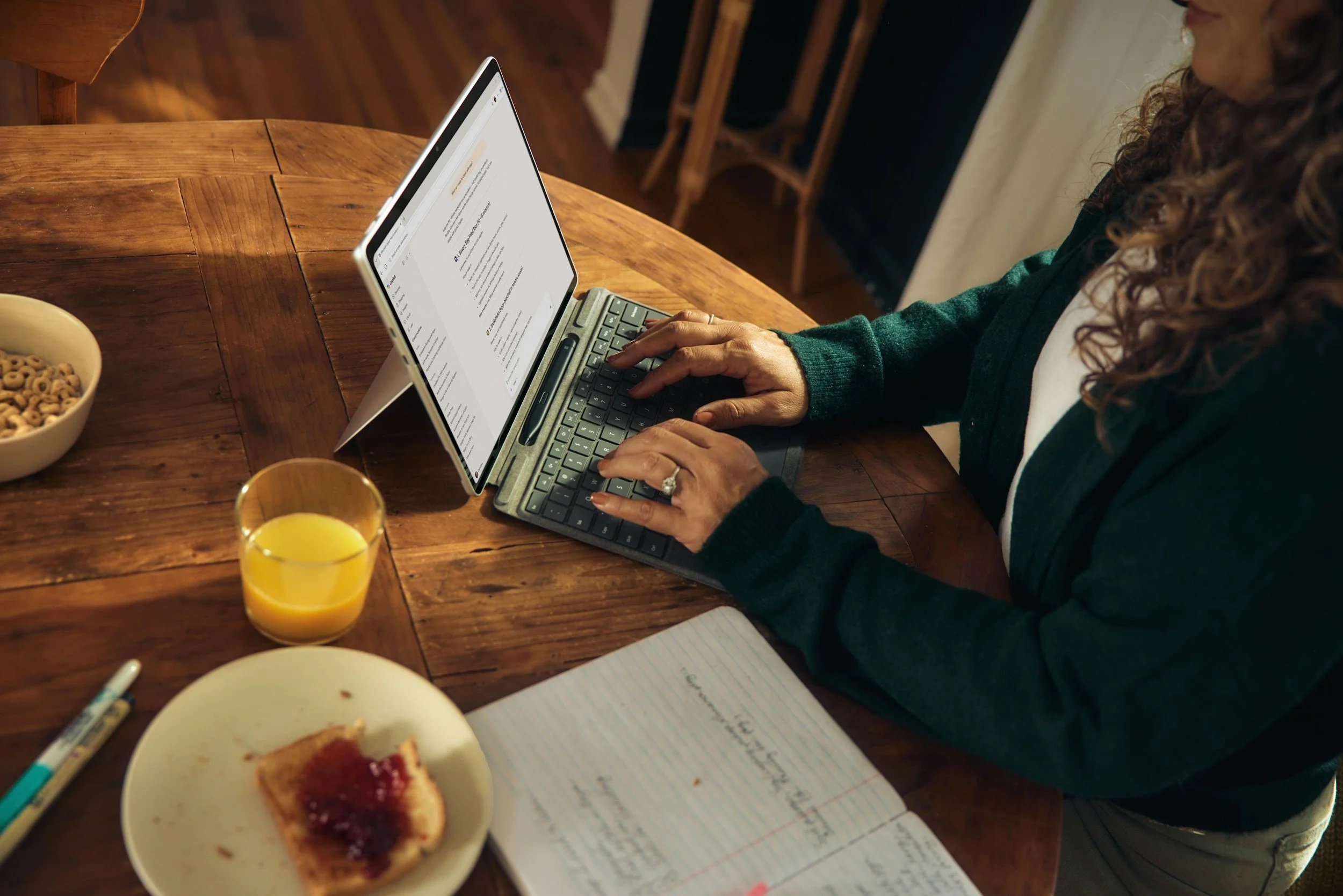 A person with curly hair wearing a dark green sweater uses a tablet with a keyboard on a wooden table, with a notebook, a glass of orange juice, a bowl of cereal, and a piece of toast with jam nearby.