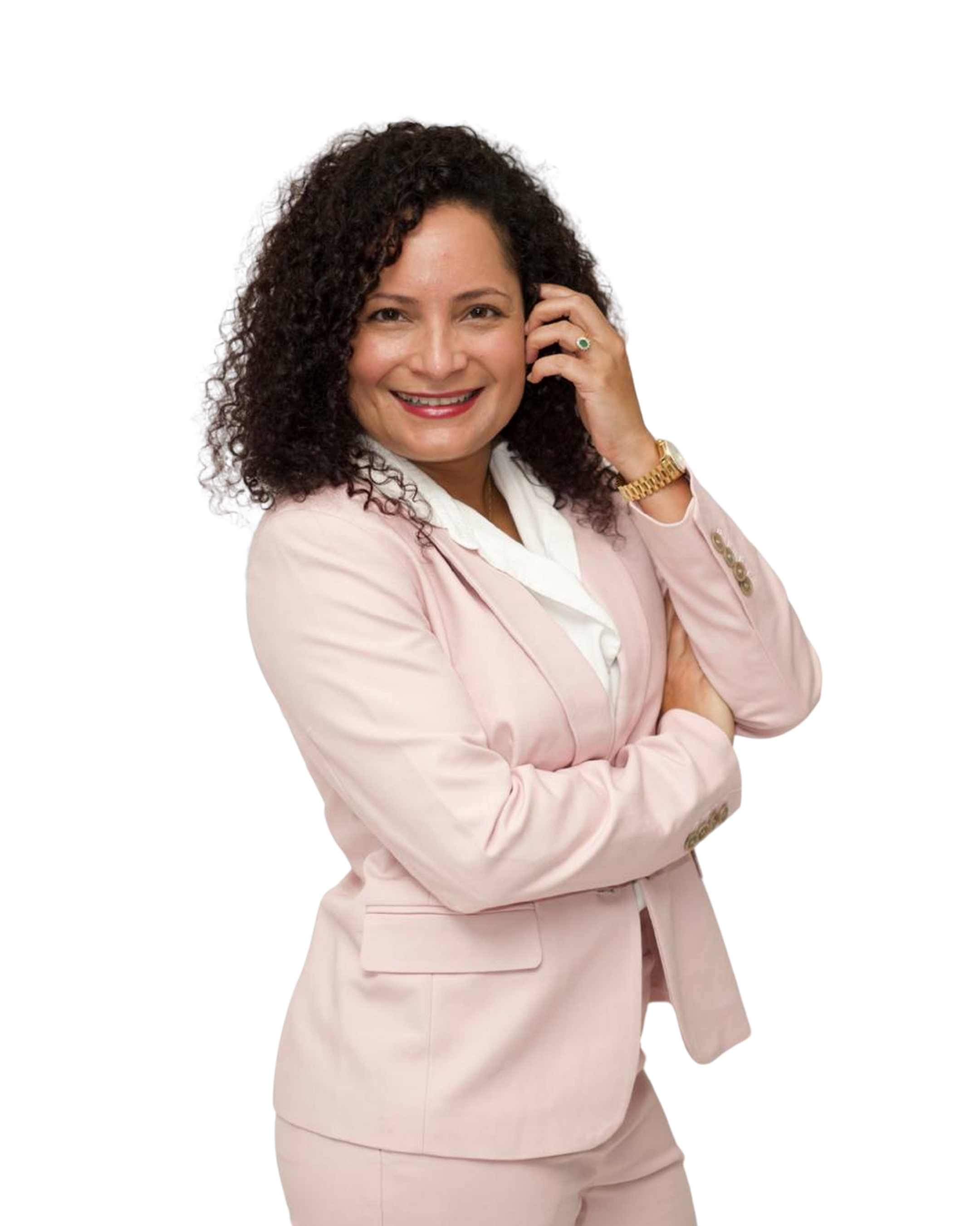 A woman with curly dark hair smiling and wearing a light pink suit with a white blouse, standing with arms crossed and touching her ear.