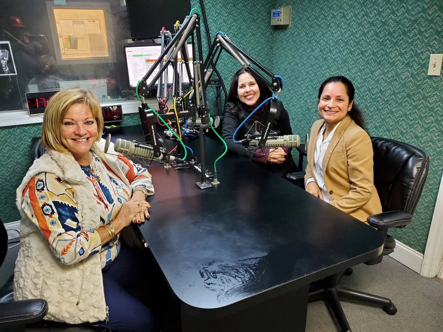 Three women sitting in a radio studio with microphones, smiling at the camera.