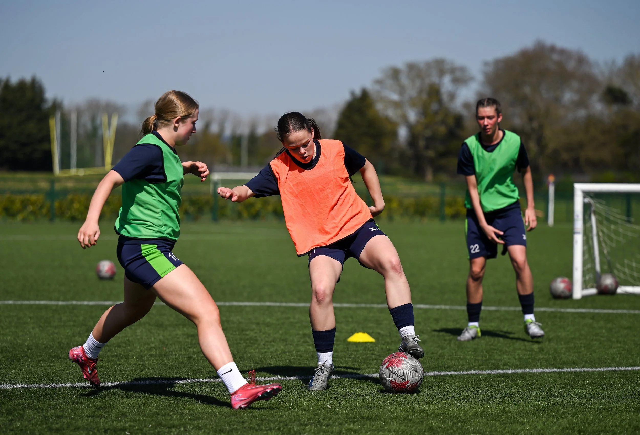 Three female soccer players practicing on the field, with two wearing green bibs and one wearing an orange bib, during daytime.