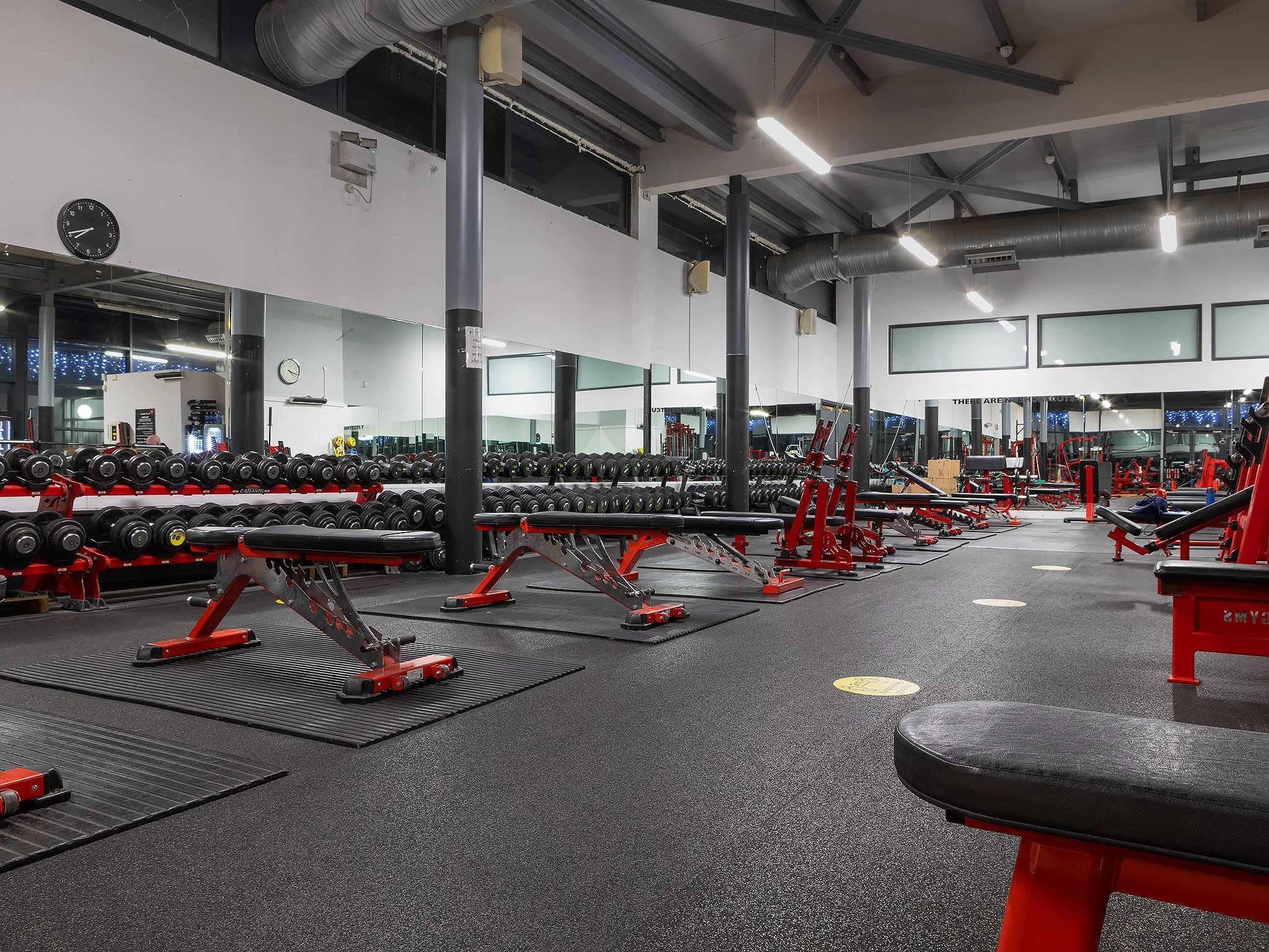 Empty gym with black rubber flooring, red and black workout equipment, benches, dumbbells, and mirrors on the wall, illuminated by ceiling lights.