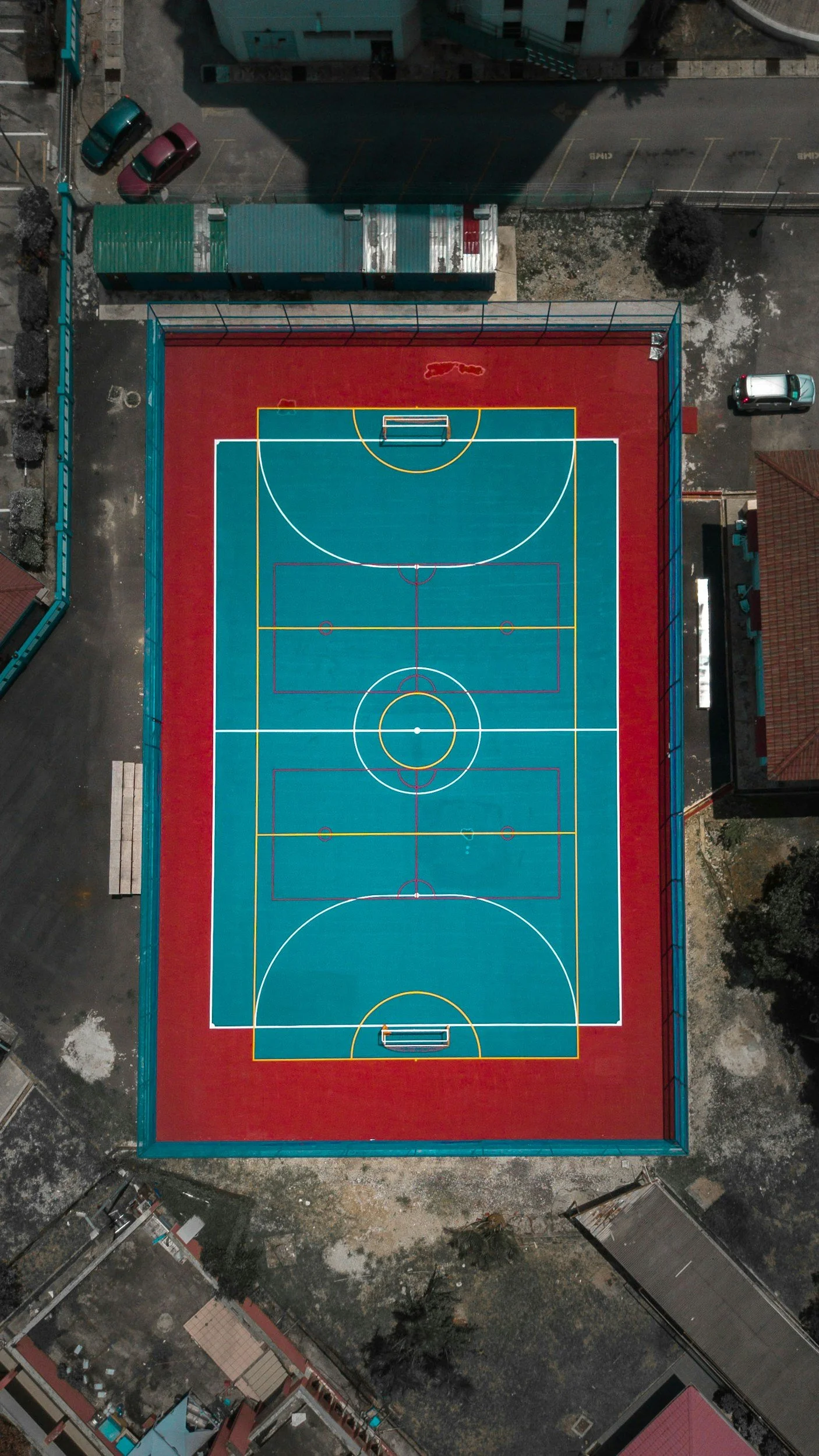 A top-down view of an outdoor multi-sport court with different colored markings for basketball and other games, enclosed by a fence, with parking and surrounding buildings.
