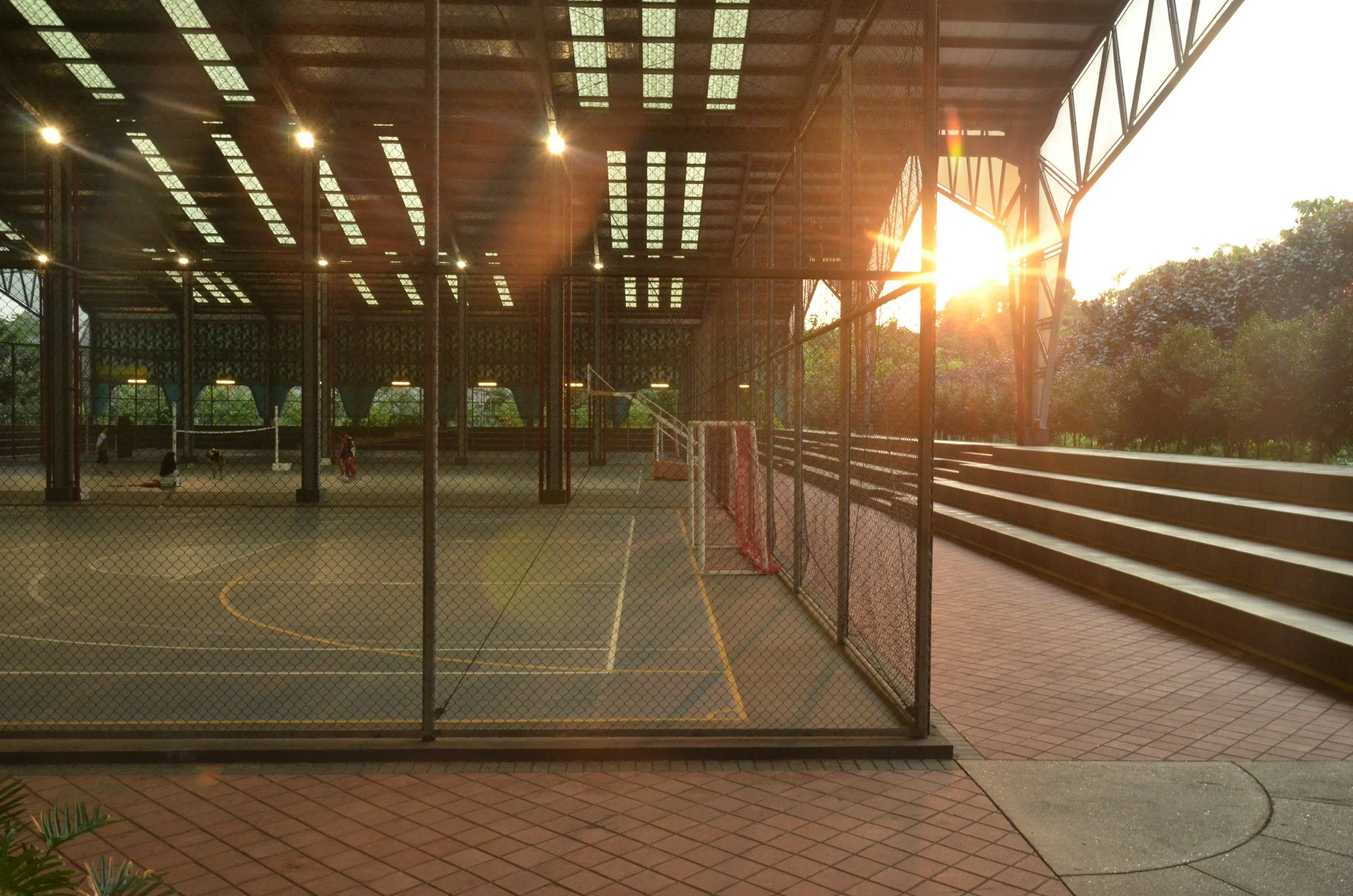 An outdoor sports court enclosed by chain-link fencing with a roof overhead. The court has lines and a small goal, and there are a few people playing. The sun is setting, casting a warm glow and creating lens flare.