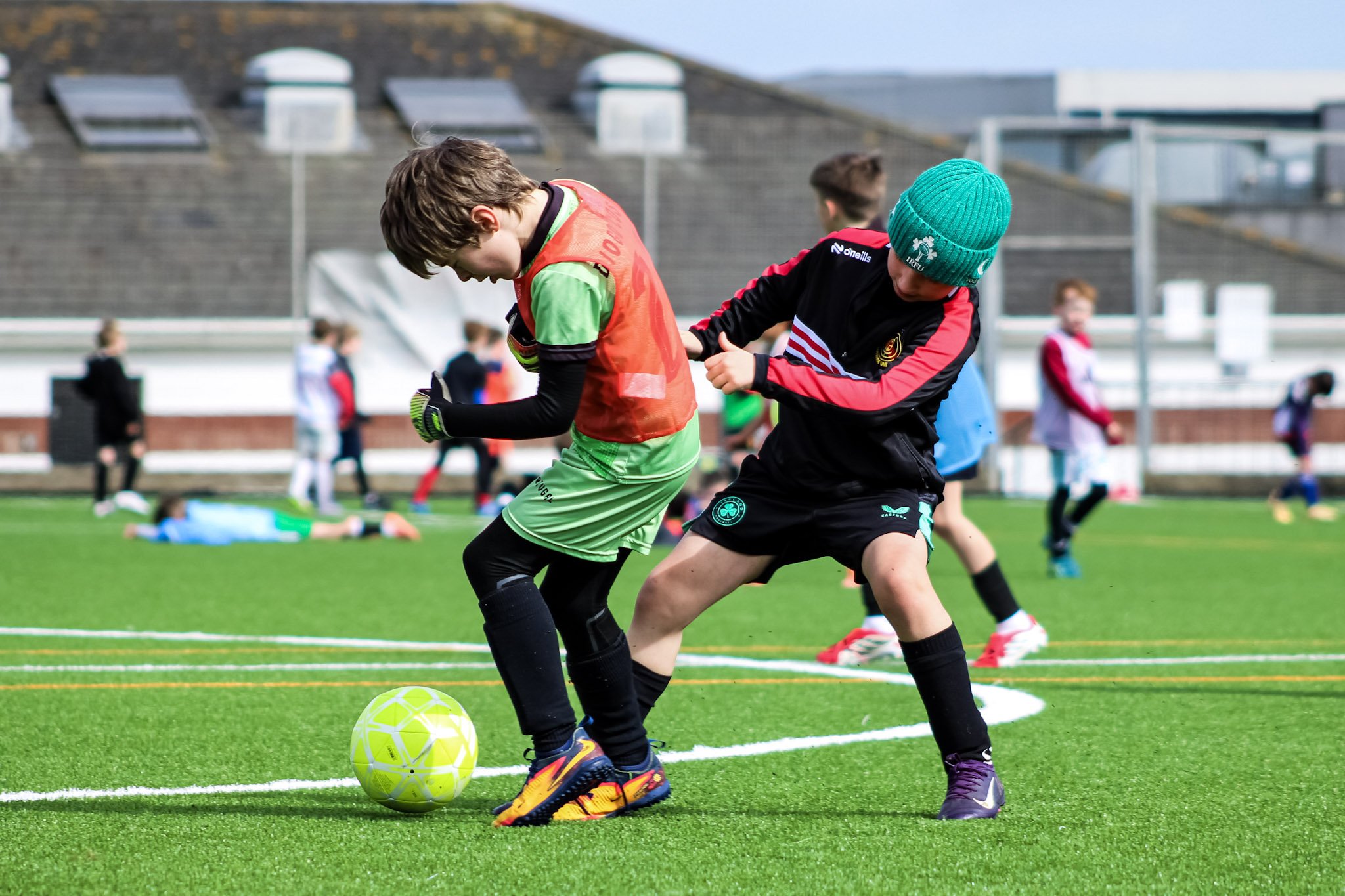 Two young boys are playing soccer on a green field. One boy wears a red and green jersey and black shorts, and the other wears a black jacket, black shorts, and a green beanie. The boy in the jersey is about to kick a bright yellow soccer ball. In the background, there are other children and a large stadium with empty seats.