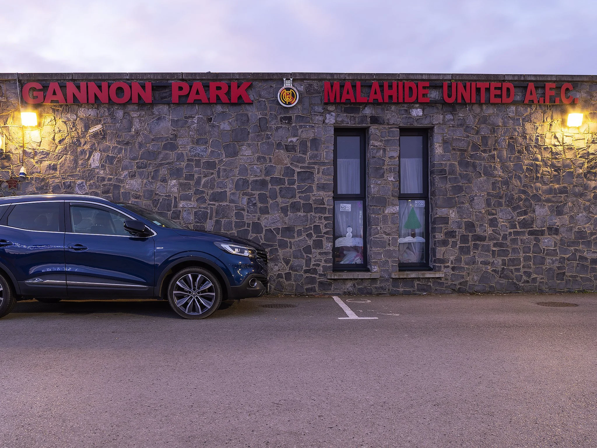 Stone building with red sign that reads 'Gannon Park Malahide United A.F.C.' and two black windows, with a parked blue car in front.