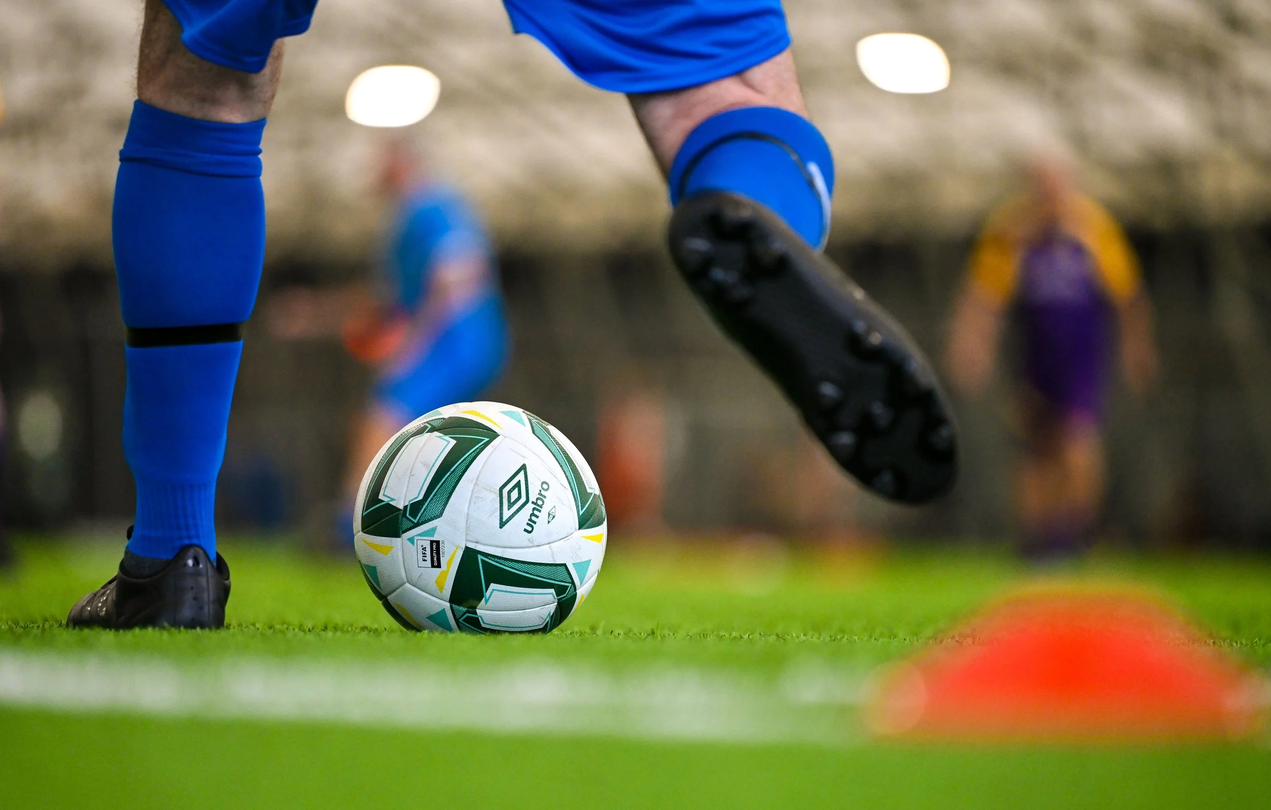 Close-up of a soccer player's legs and feet about to kick a white and green soccer ball on a field, with orange cones in the foreground and people blurred in the background.